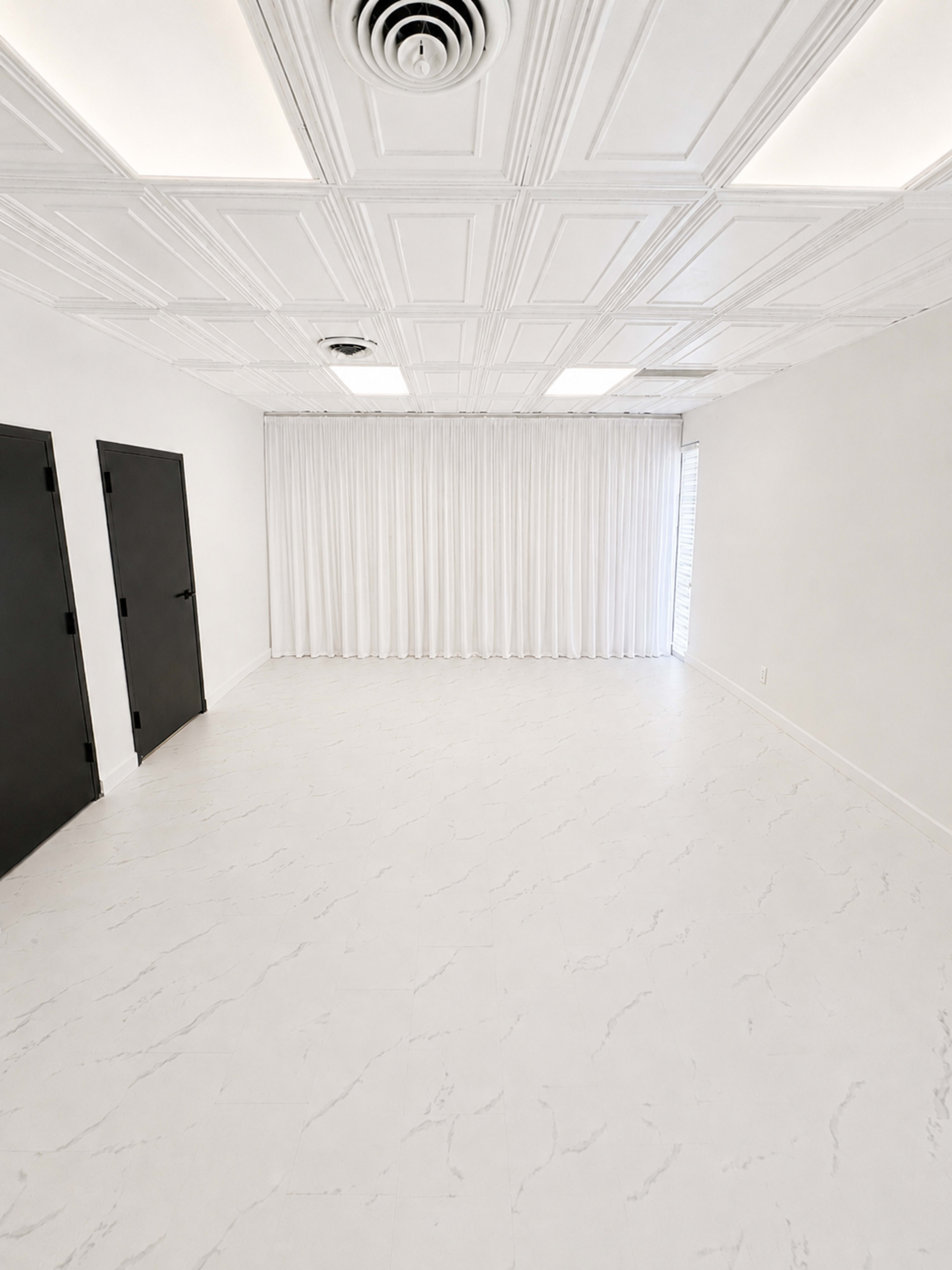 The image shows an empty, white room with two black doors and a large curtain along one wall.