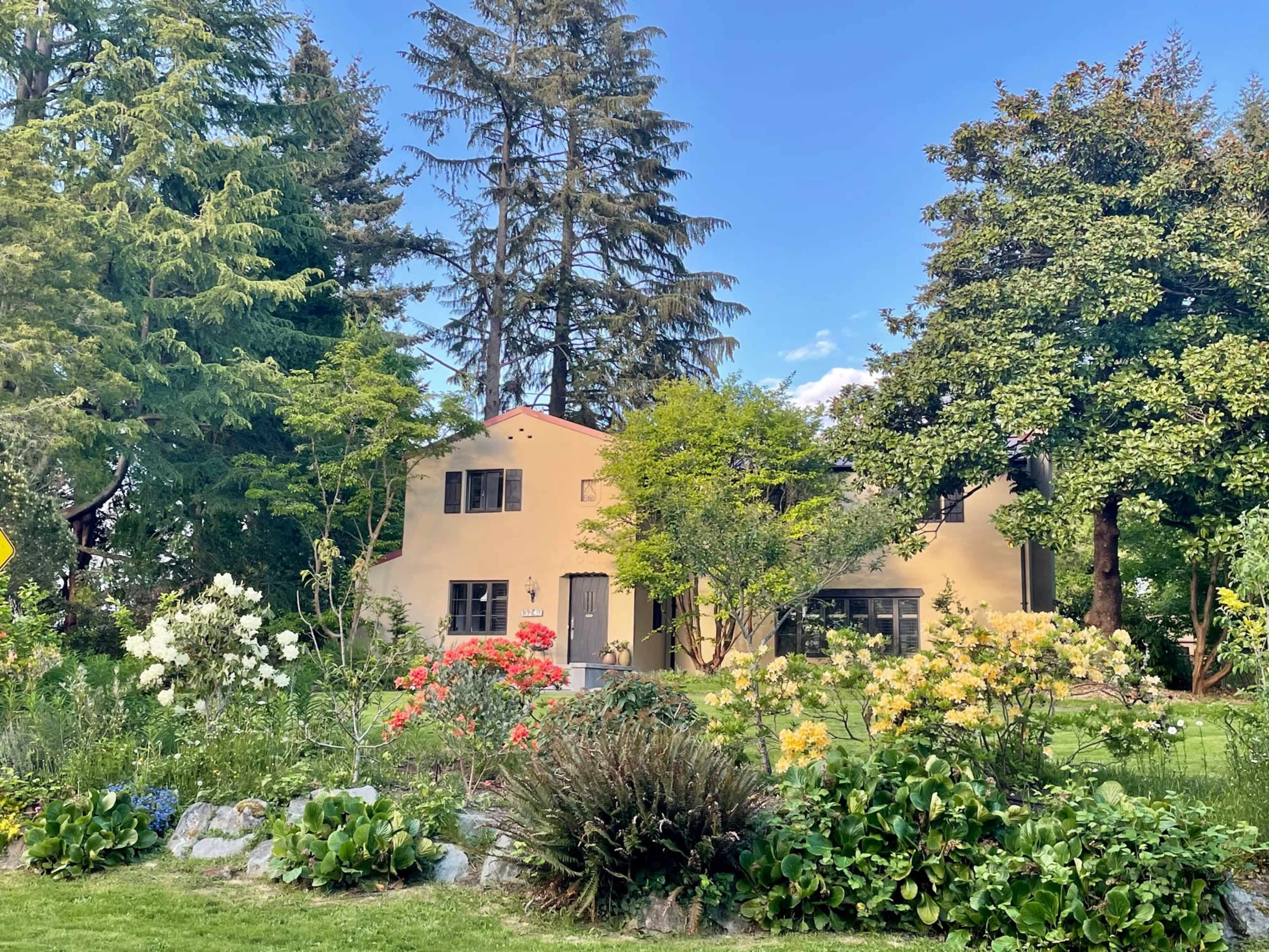 A two-story house with a light-colored exterior is surrounded by colorful flower gardens and tall trees.