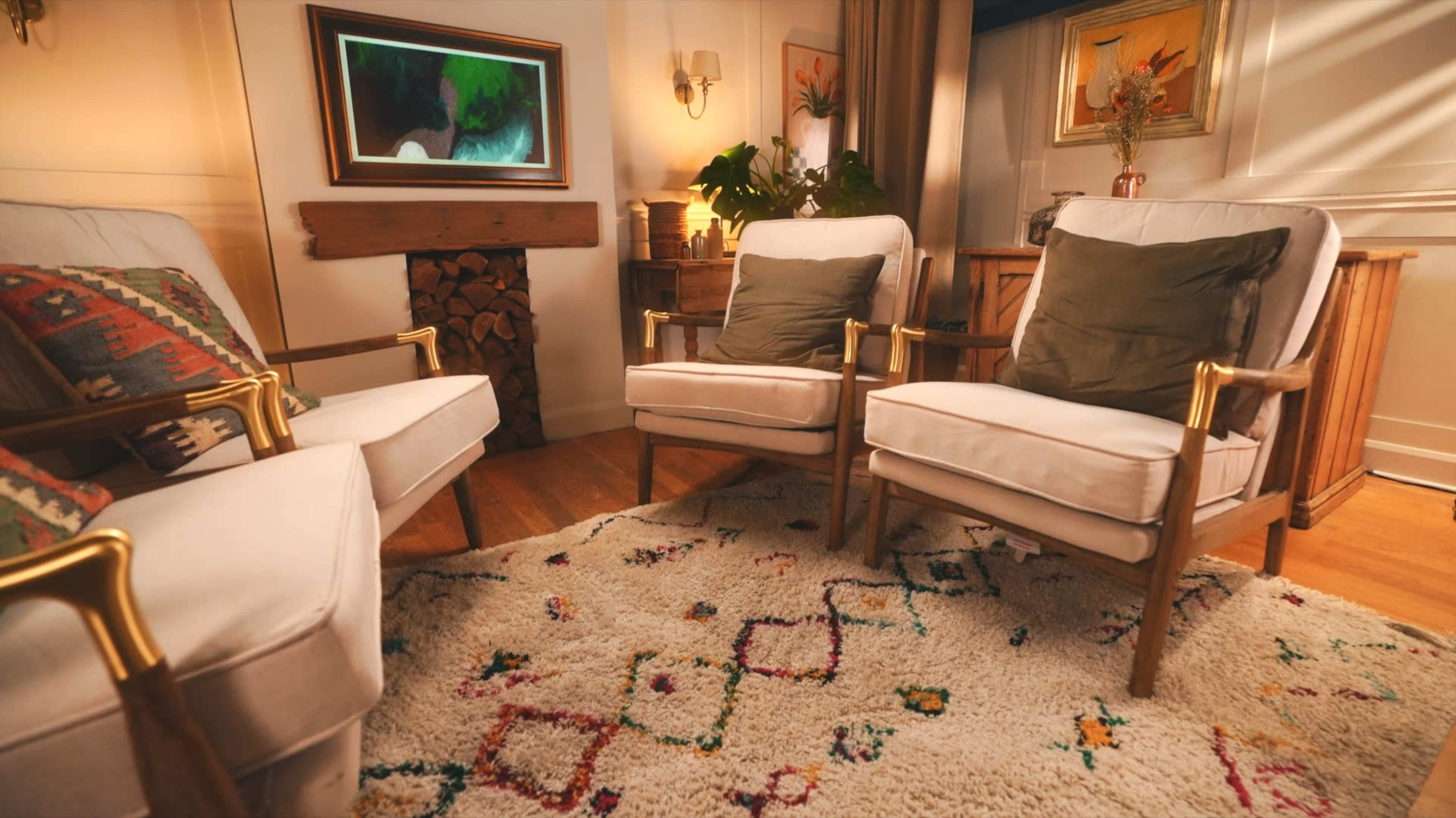 The image features a cozy living room with three upholstered chairs arranged around a decorative rug, a small wooden side table, and a stack of logs near the wall.