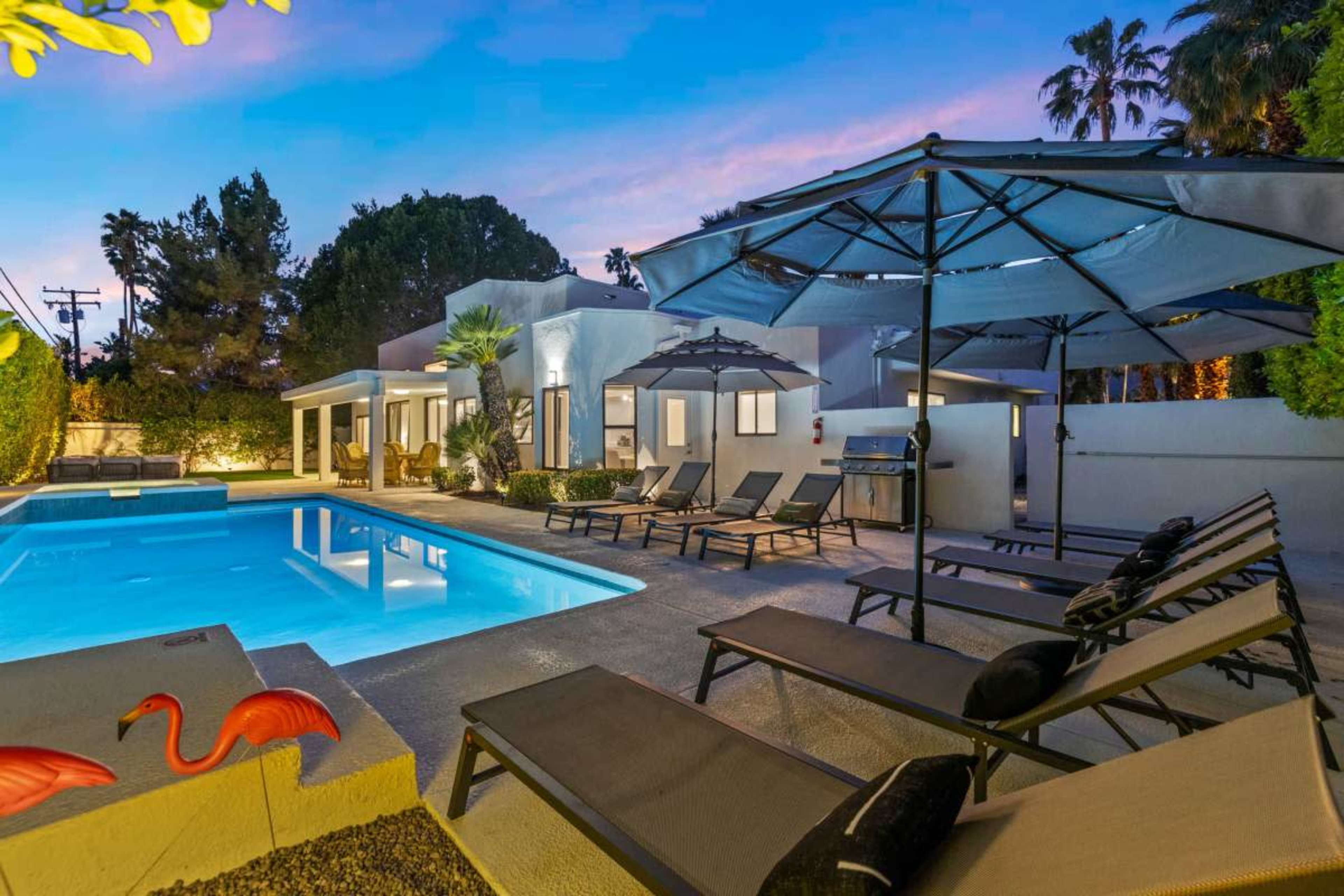 A modern patio area features a swimming pool surrounded by lounge chairs and umbrellas, set against a backdrop of palm trees at dusk.