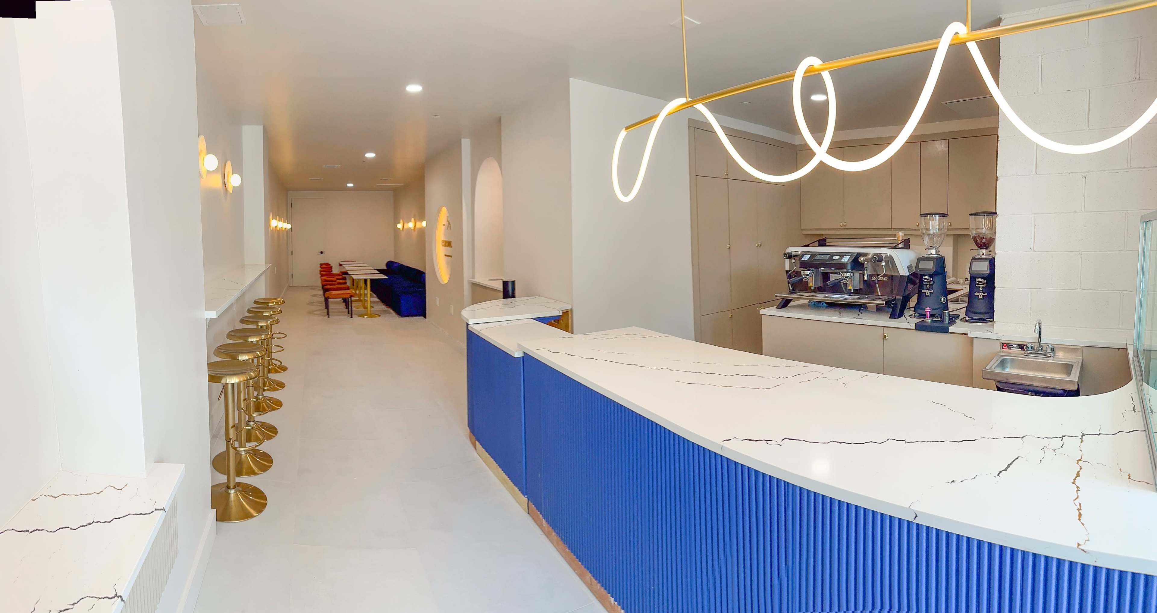 The image shows a modern cafe interior featuring a blue and white patterned counter, seating along one side, and a coffee machine in the back.