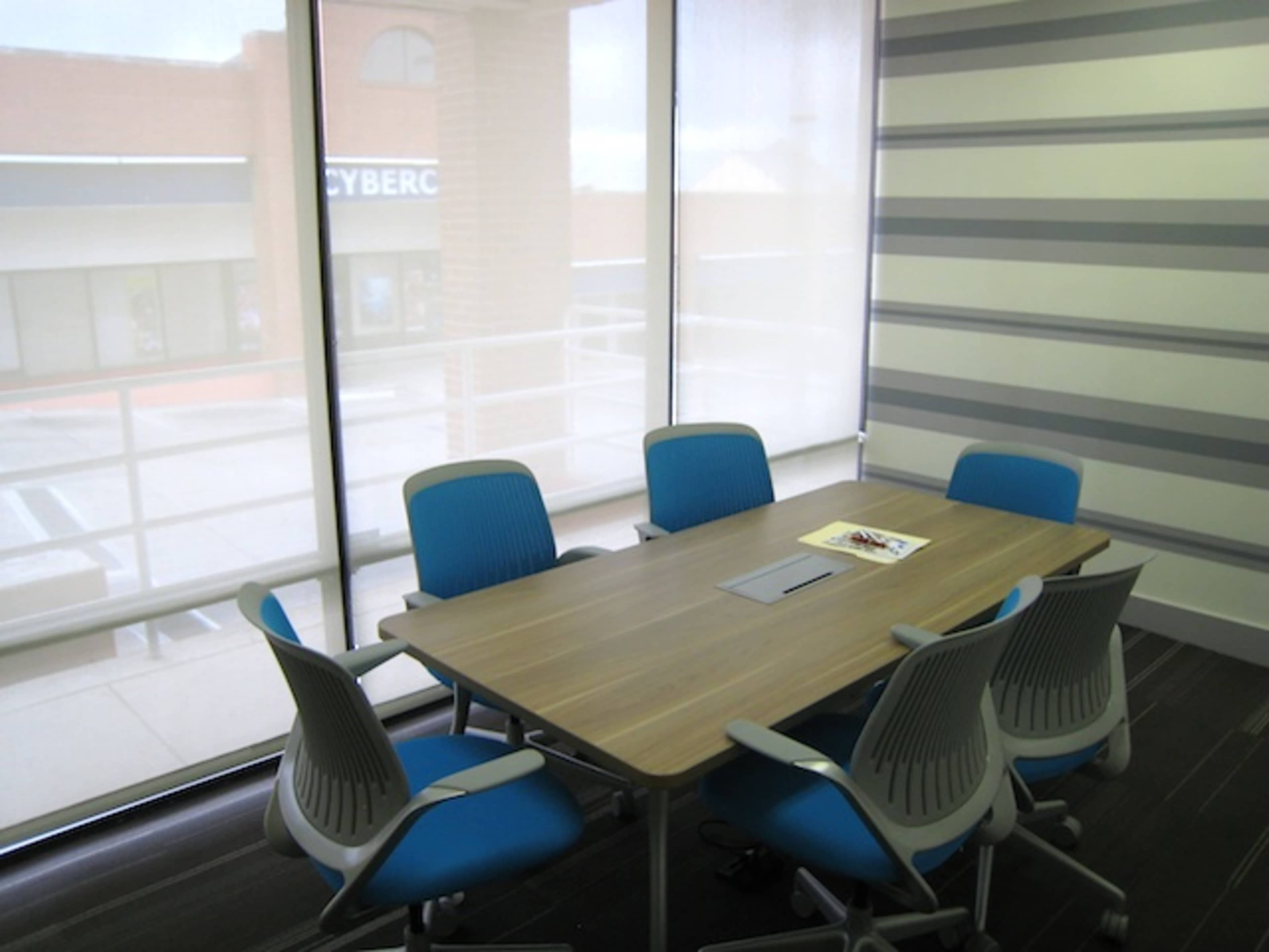 A conference room features a wooden table surrounded by six blue chairs, with large windows allowing natural light to filter through sheer blinds.