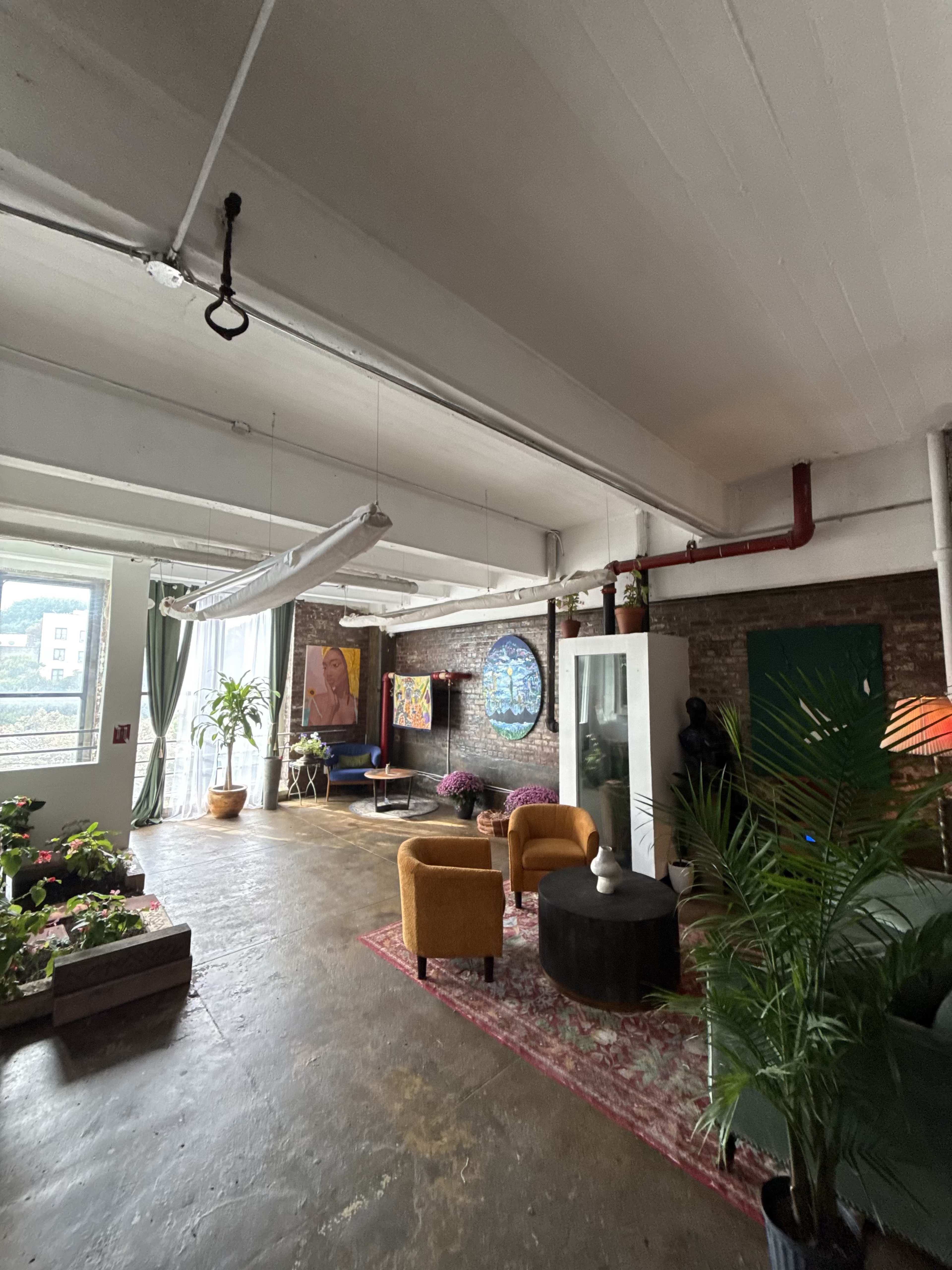 The image shows an industrial-style room with exposed brick walls, a large window, various plants, and a mix of modern furniture including yellow chairs and a round coffee table.