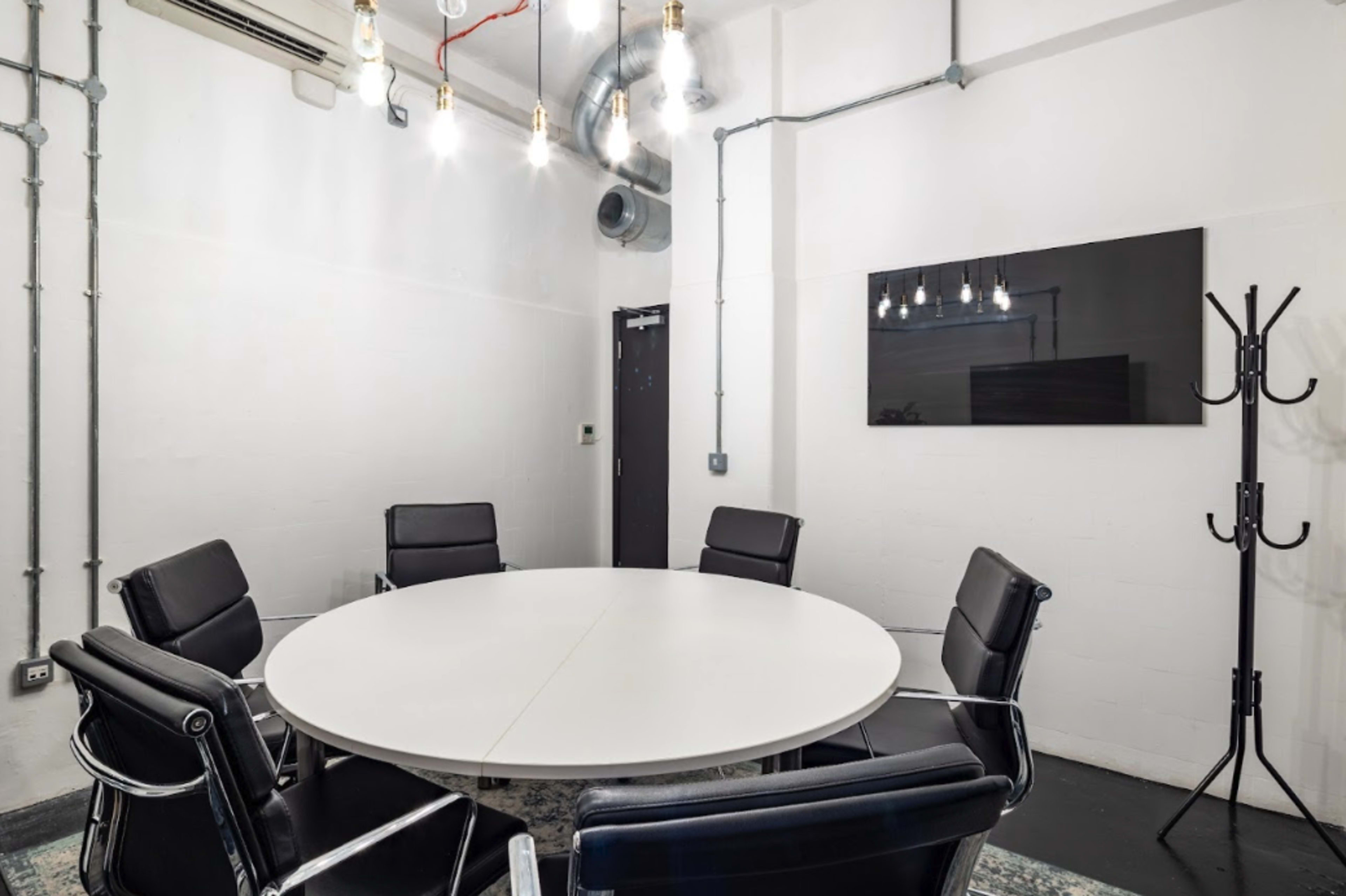 A round white table surrounded by black chairs in a minimalist meeting room with exposed pipes and overhead lights.