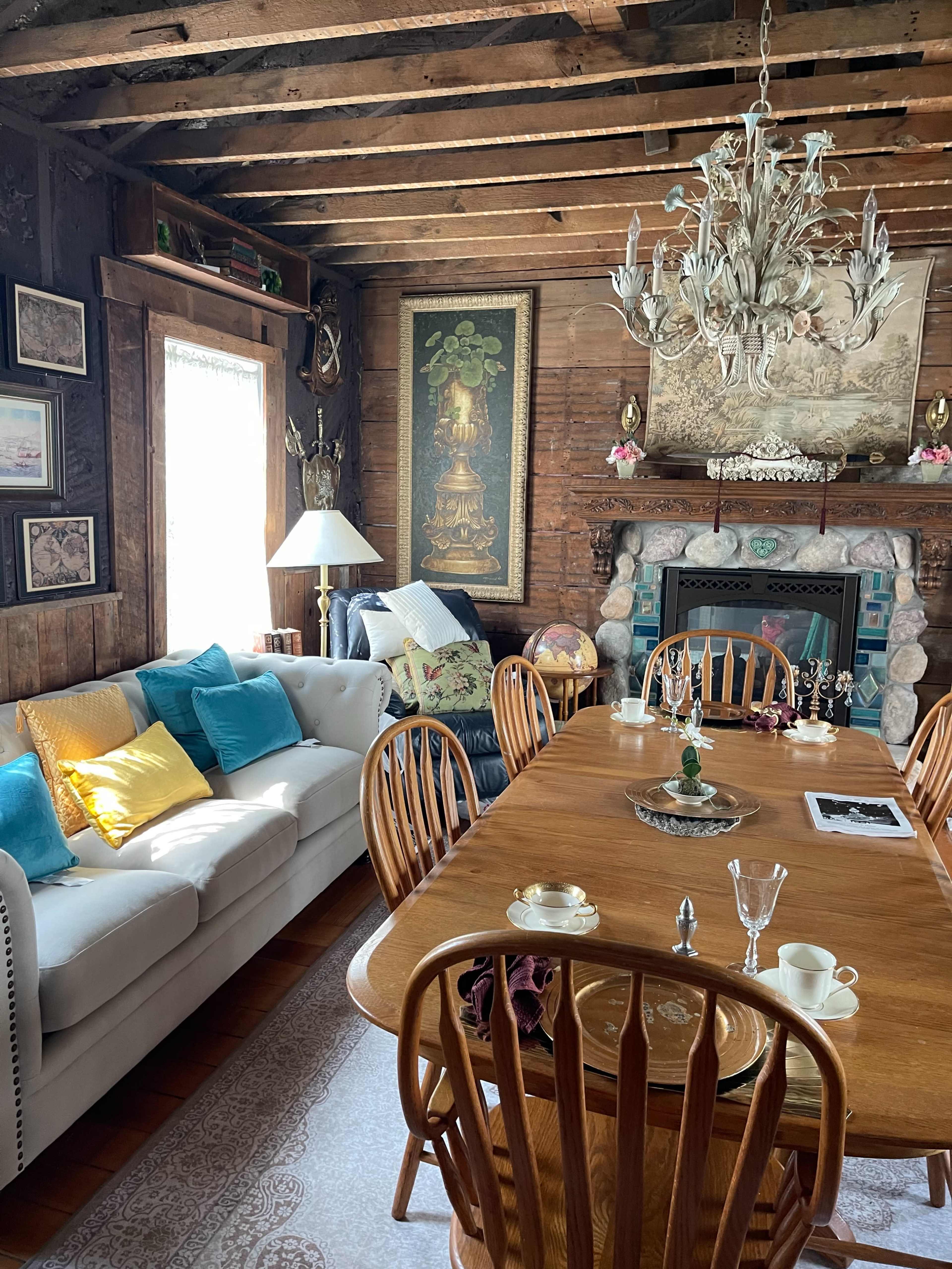 A rustic dining area with a wooden table surrounded by chairs, a white sofa adorned with colorful cushions, and various decorations on the walls and shelves.