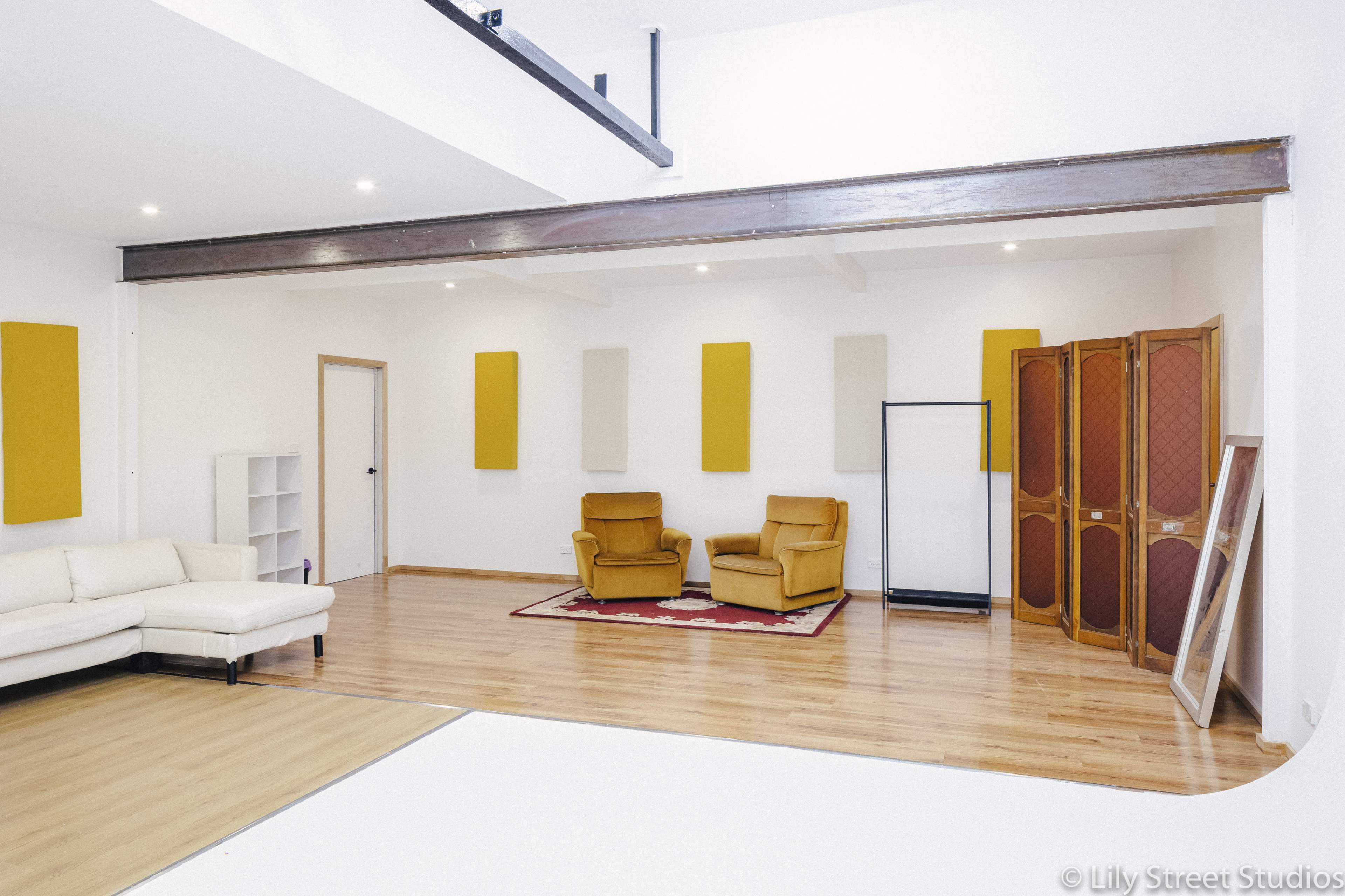 The image shows a spacious studio with a light wooden floor, a beige sofa, two yellow chairs, and decorative acoustic panels on the walls.