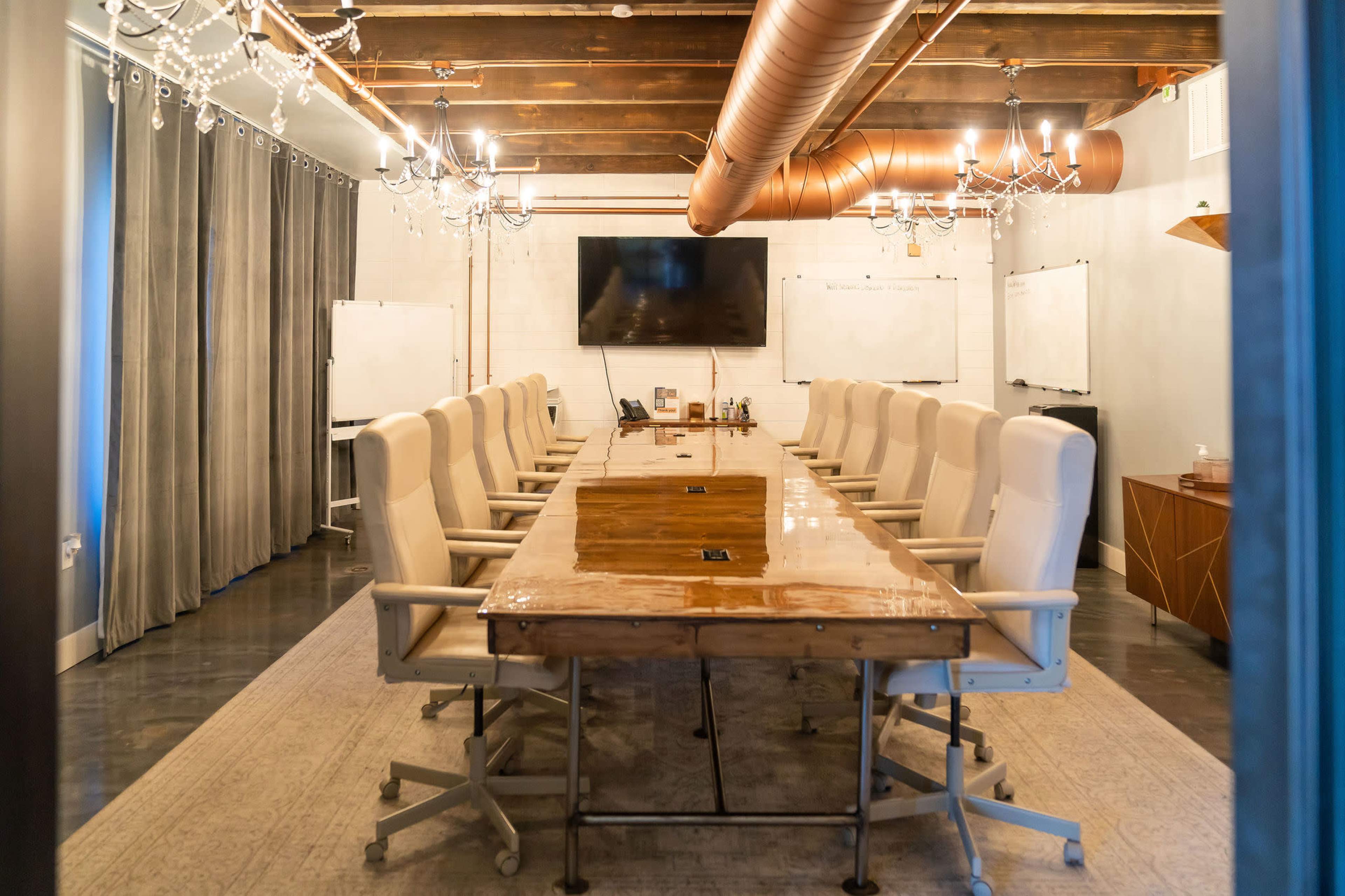 A conference room features a long wooden table surrounded by chairs, with a large screen on the wall and whiteboards on either side.