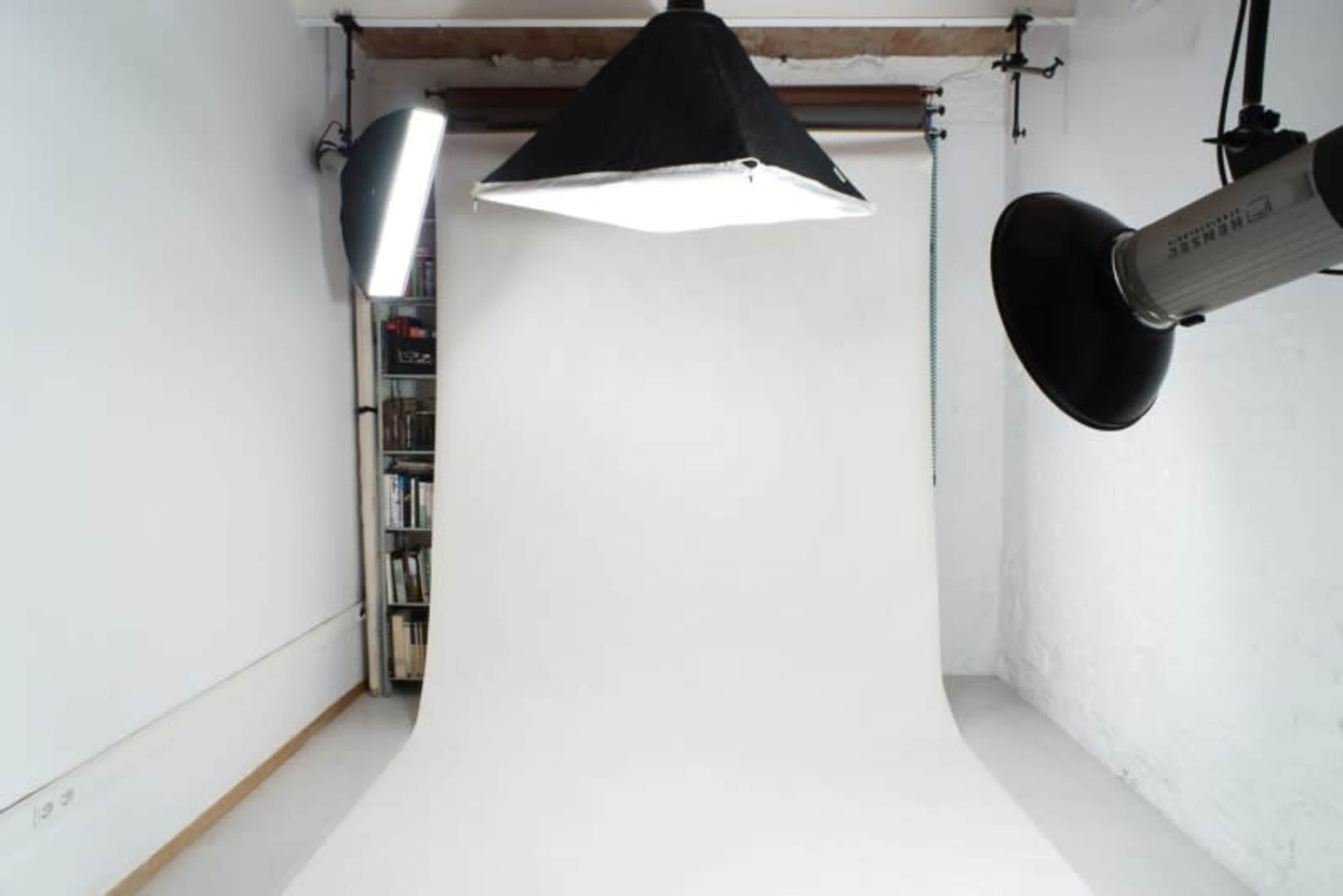 A photography studio with a white backdrop, overhead lighting, and a bookshelf in the background.