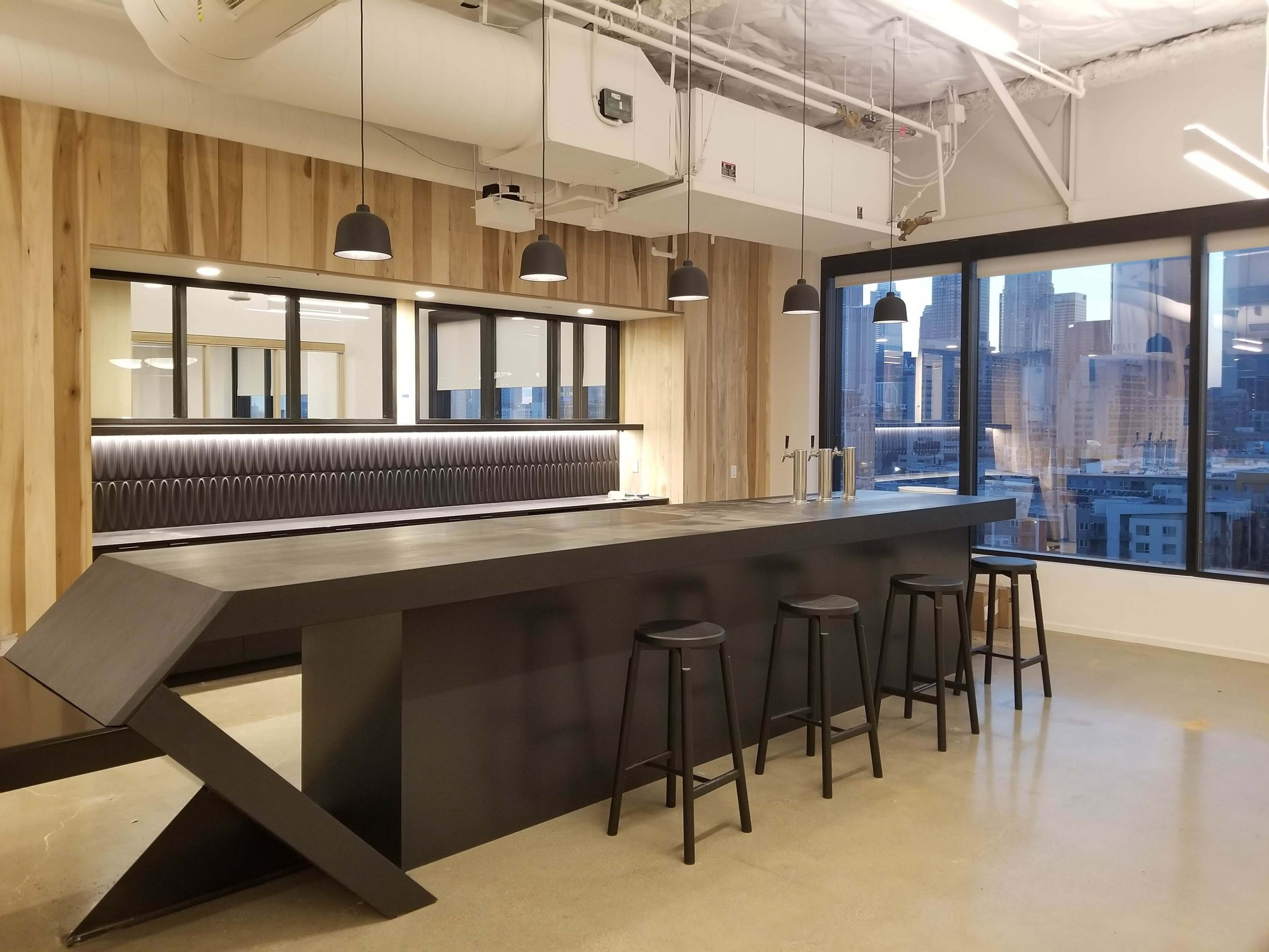 A modern office kitchen area with a black angular counter, bar stools, and large windows overlooking a cityscape.