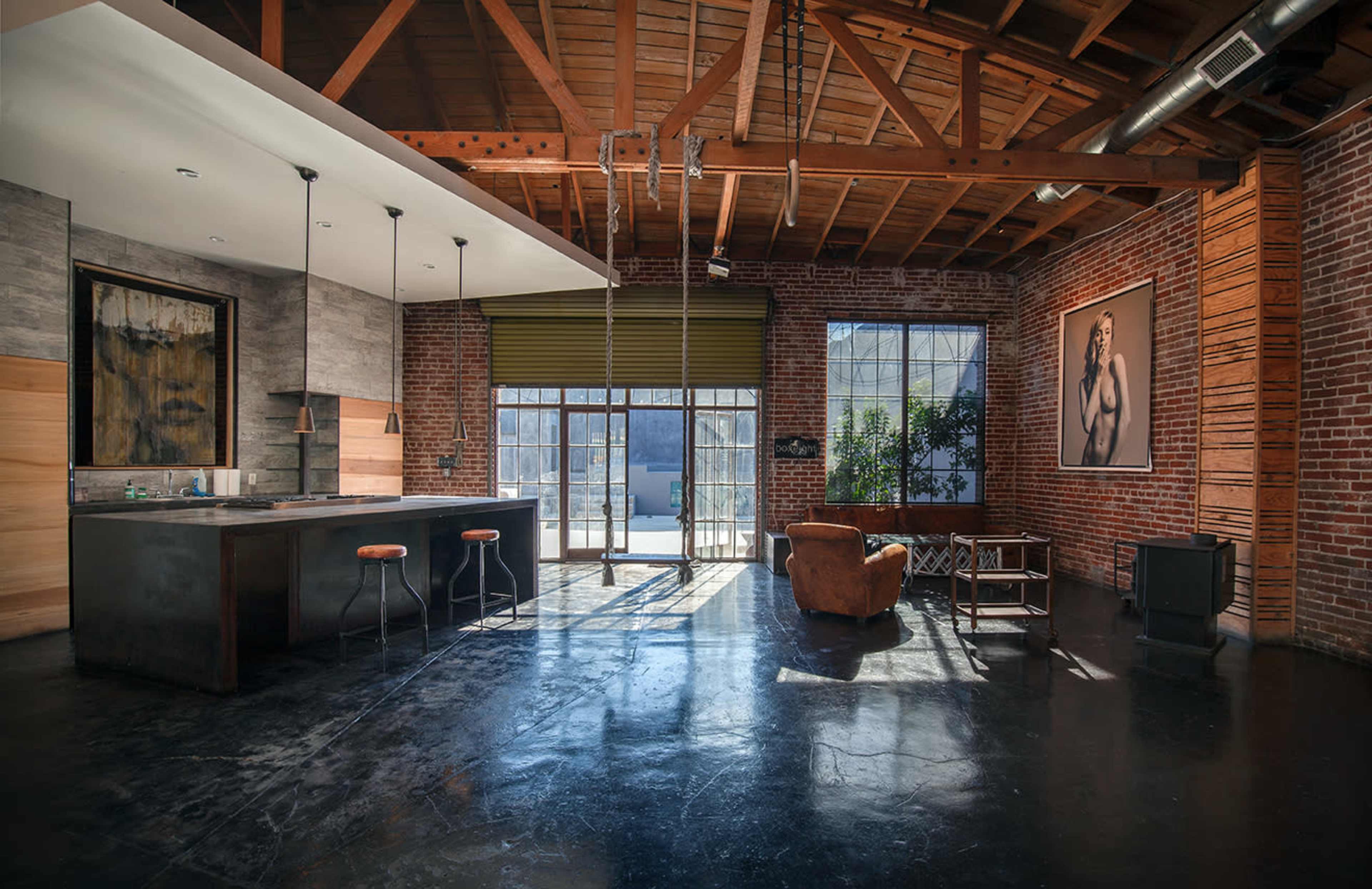 A spacious loft with exposed brick walls, industrial-style beams, a kitchen area with a dark countertop, and large windows that open to an outdoor space.