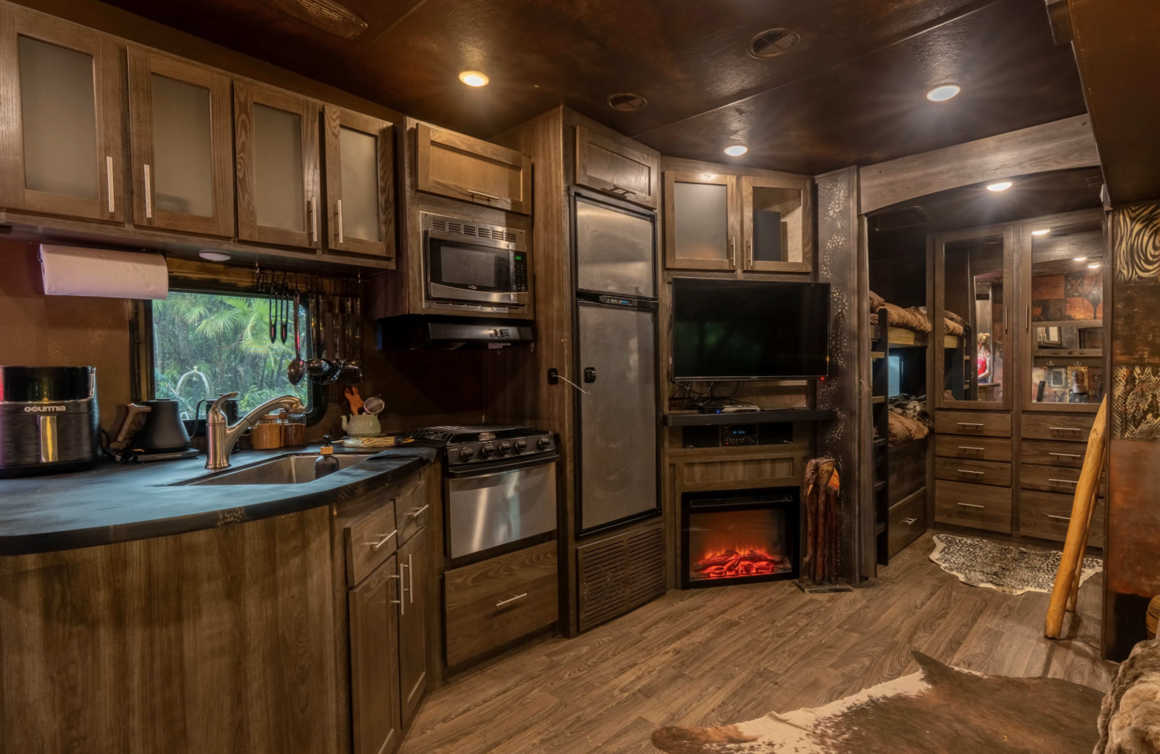 The image shows a compact kitchen area with dark wood cabinetry, stainless steel appliances, and a cozy electric fireplace in an RV interior.