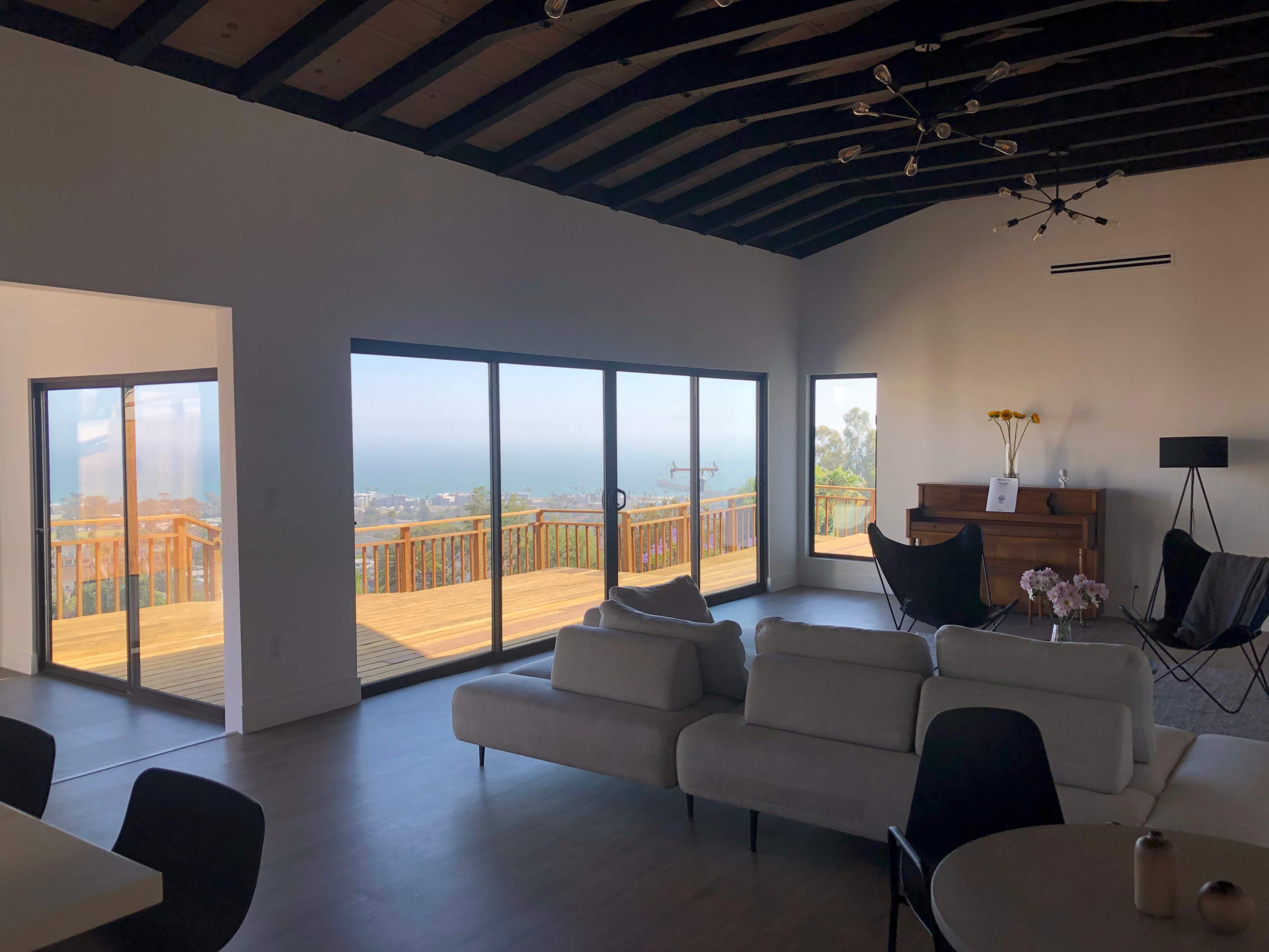 A spacious living room with large glass doors leading to a deck that overlooks a coastal view, featuring modern furniture and a wooden piano.