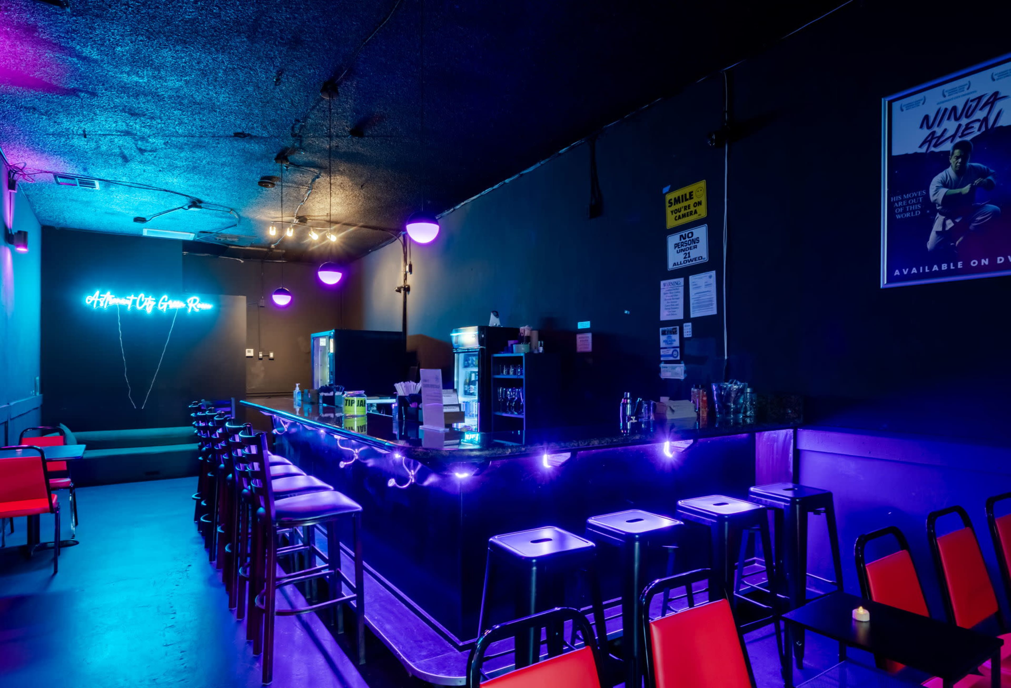 A dimly lit bar features a long counter with purple neon lighting and red chairs arranged around it.