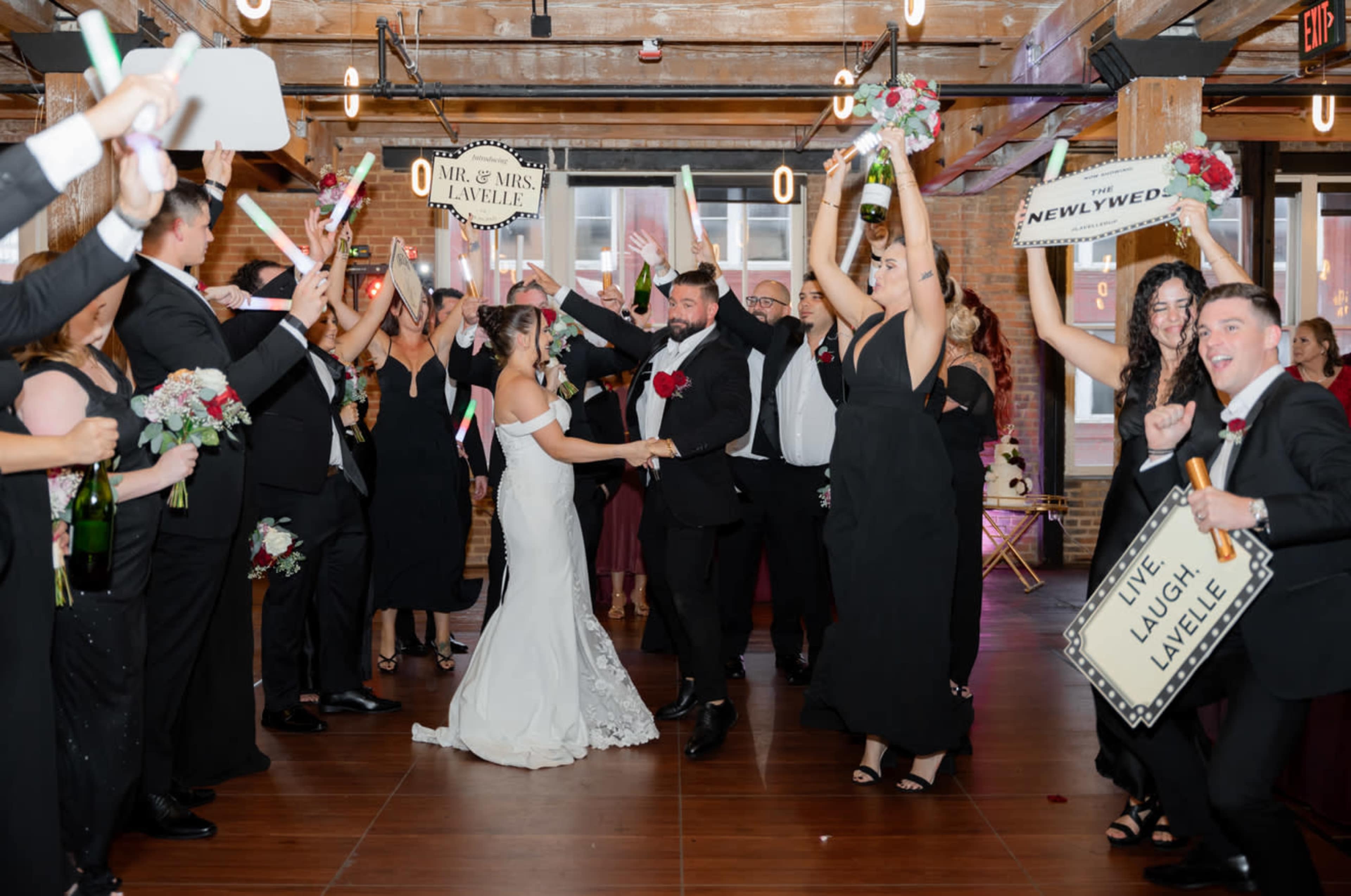 A bride and groom joyfully exit their wedding celebration while guests hold up signs and bottles of champagne in a decorated venue.