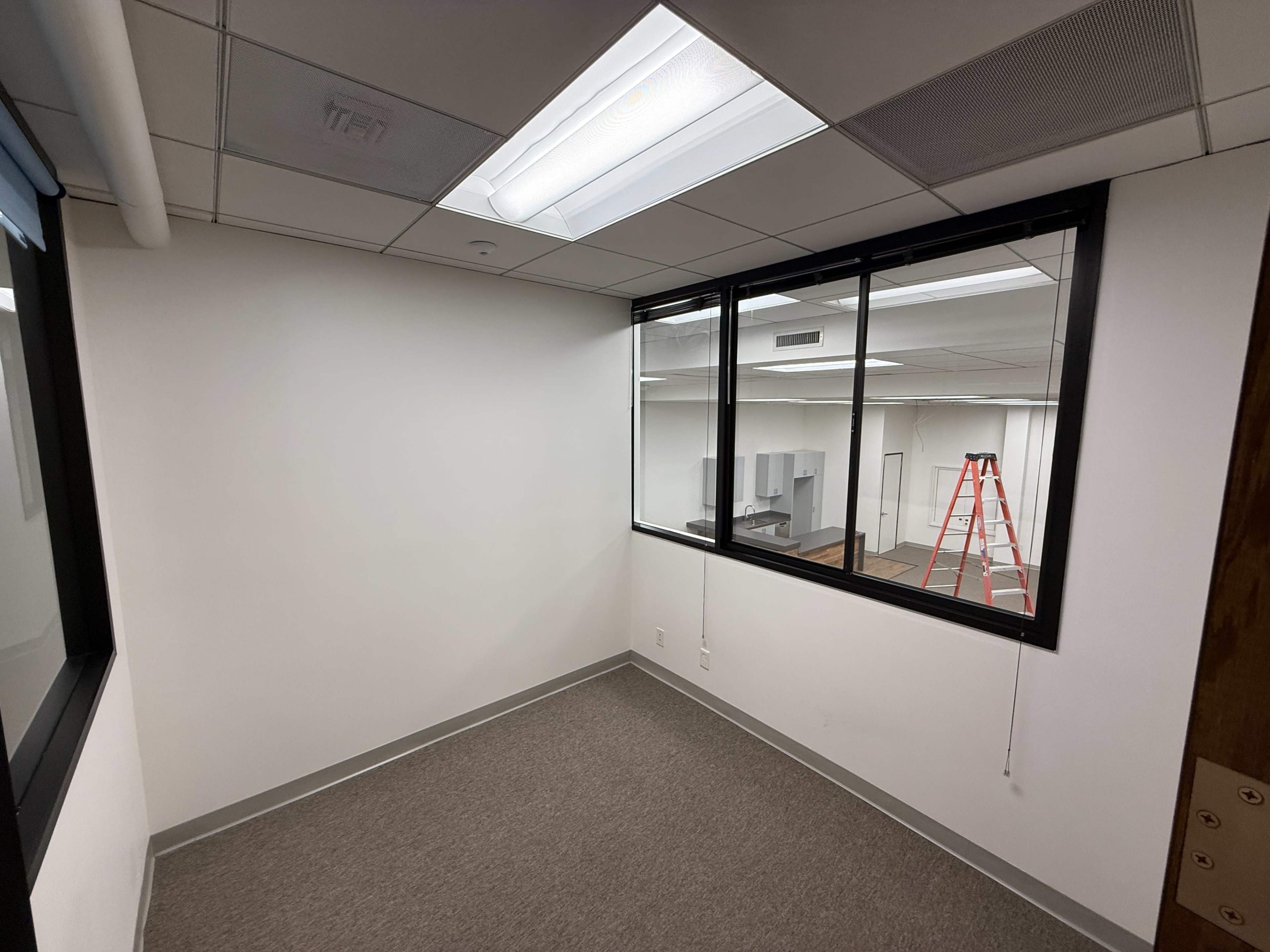 The image shows an empty office space with a gray carpet, white walls, a large window, and a ladder in the background.