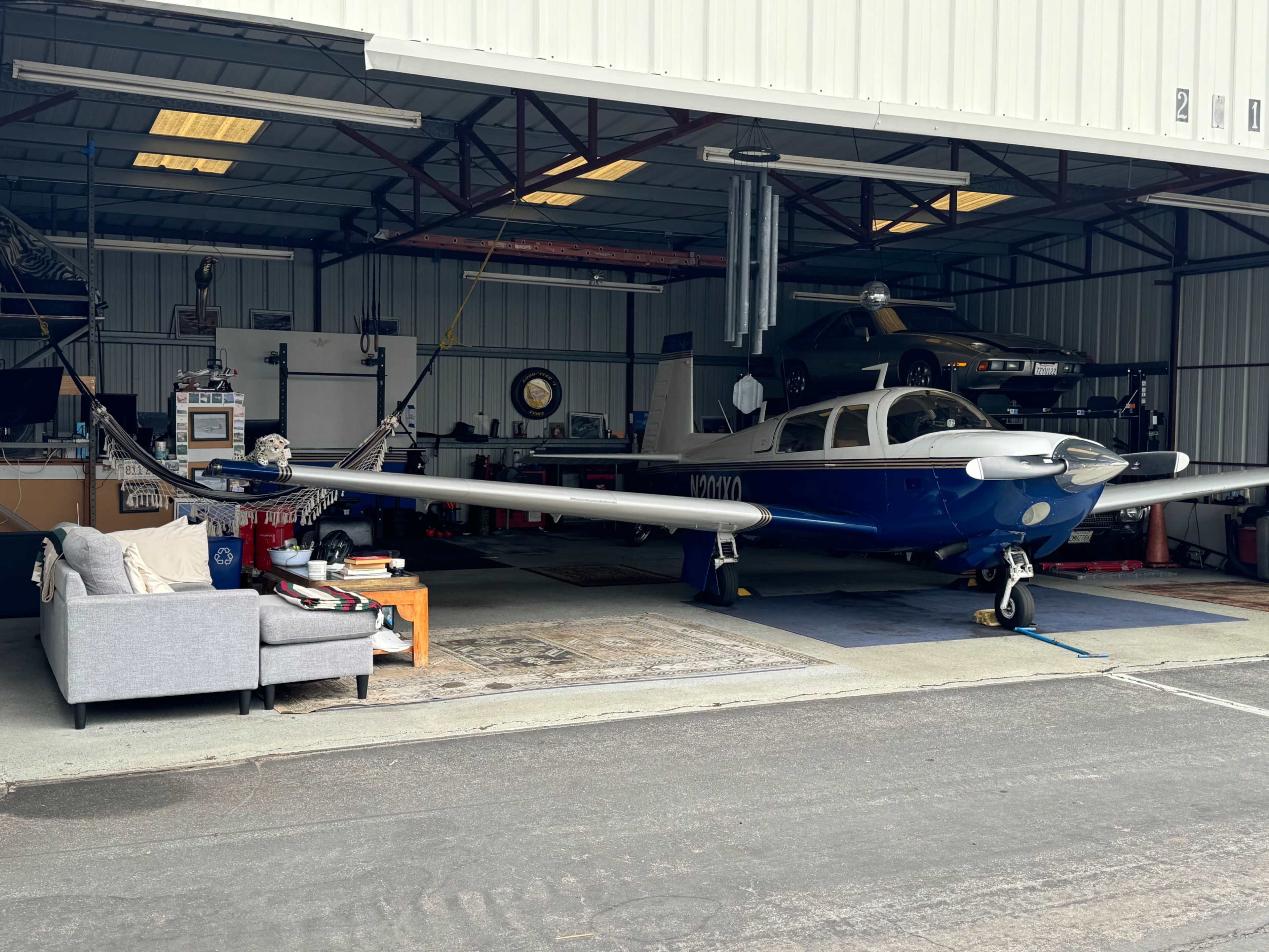 A blue aircraft is parked inside a hangar alongside a black car and various furniture.