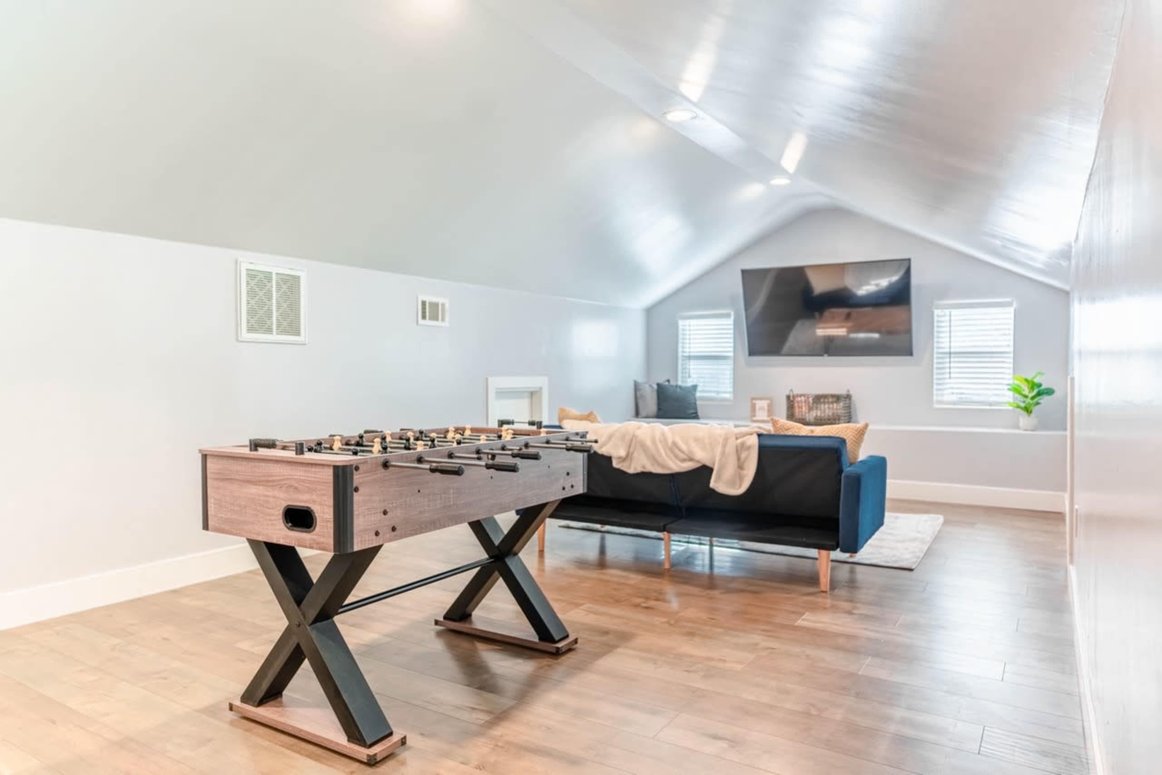 A spacious, well-lit attic features a foosball table, a sofa, and a wall-mounted television.