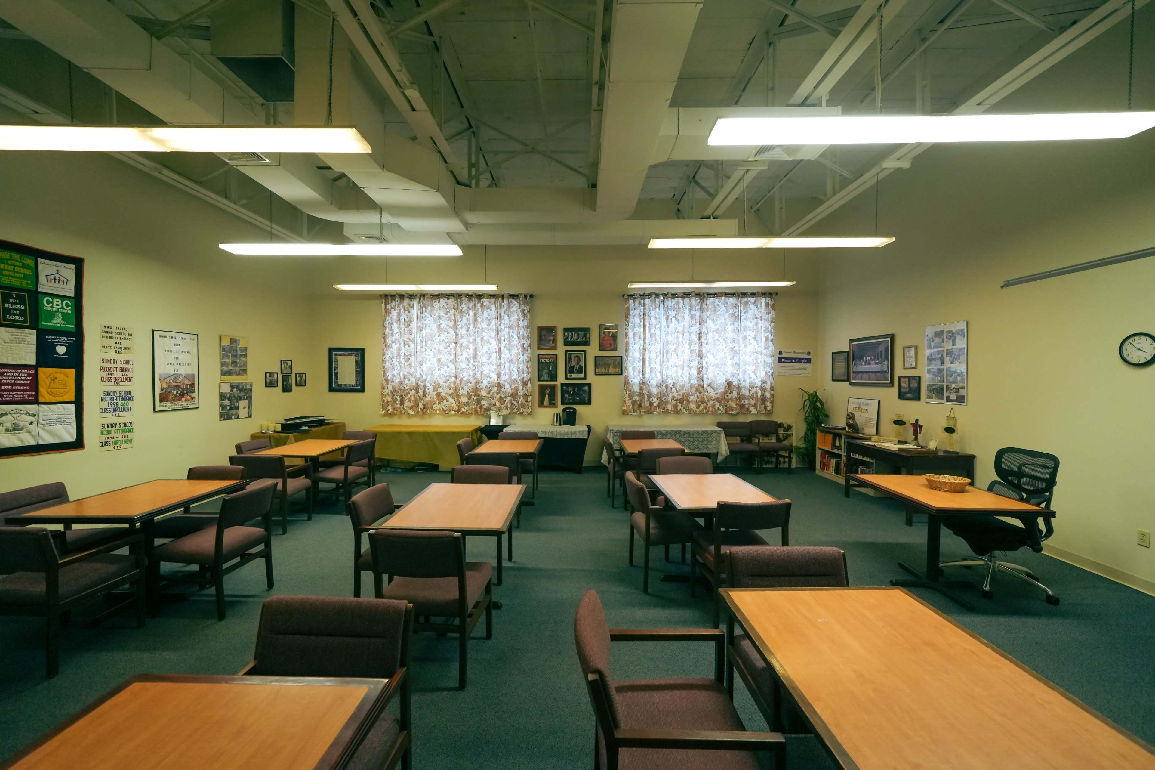 A spacious room features several tables and chairs arranged in a community center setting, with large windows dressed in floral curtains.