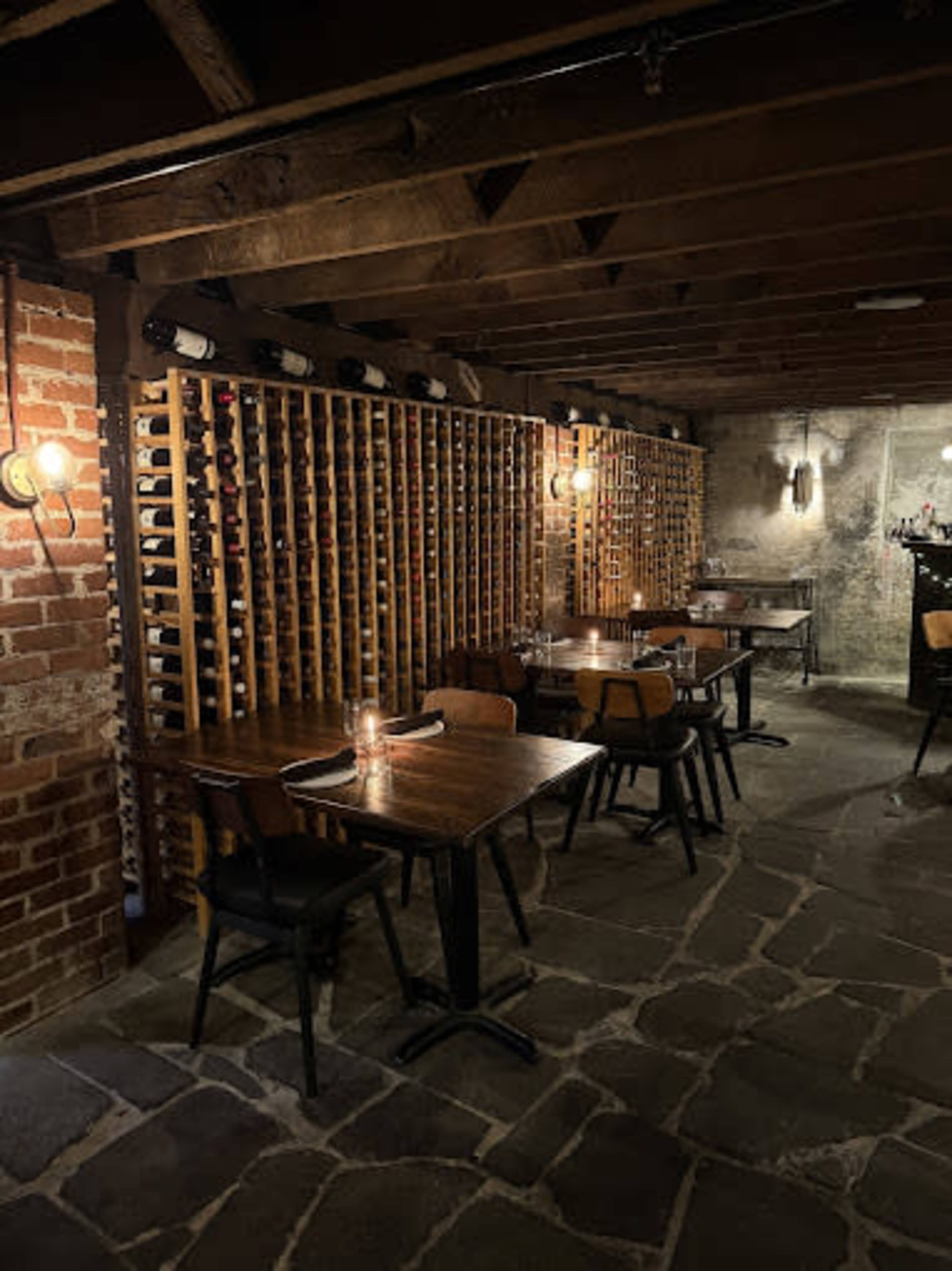 A dimly lit restaurant cellar features wooden tables and chairs arranged beside wine racks filled with bottles, set against exposed brick and stone walls.