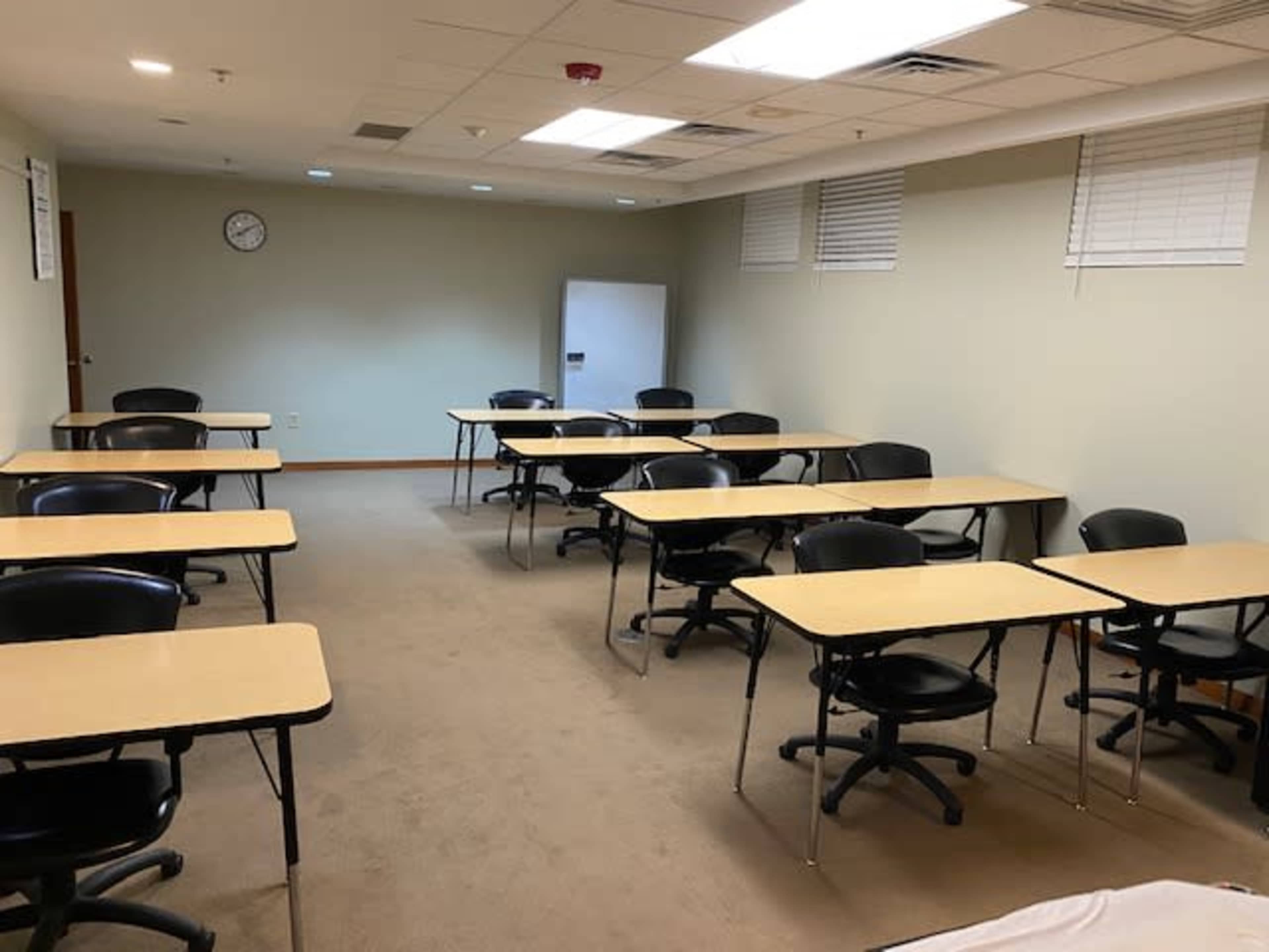 A classroom with several rows of tables and chairs arranged neatly on a carpeted floor, along with a wall clock and a refrigerator in the background.