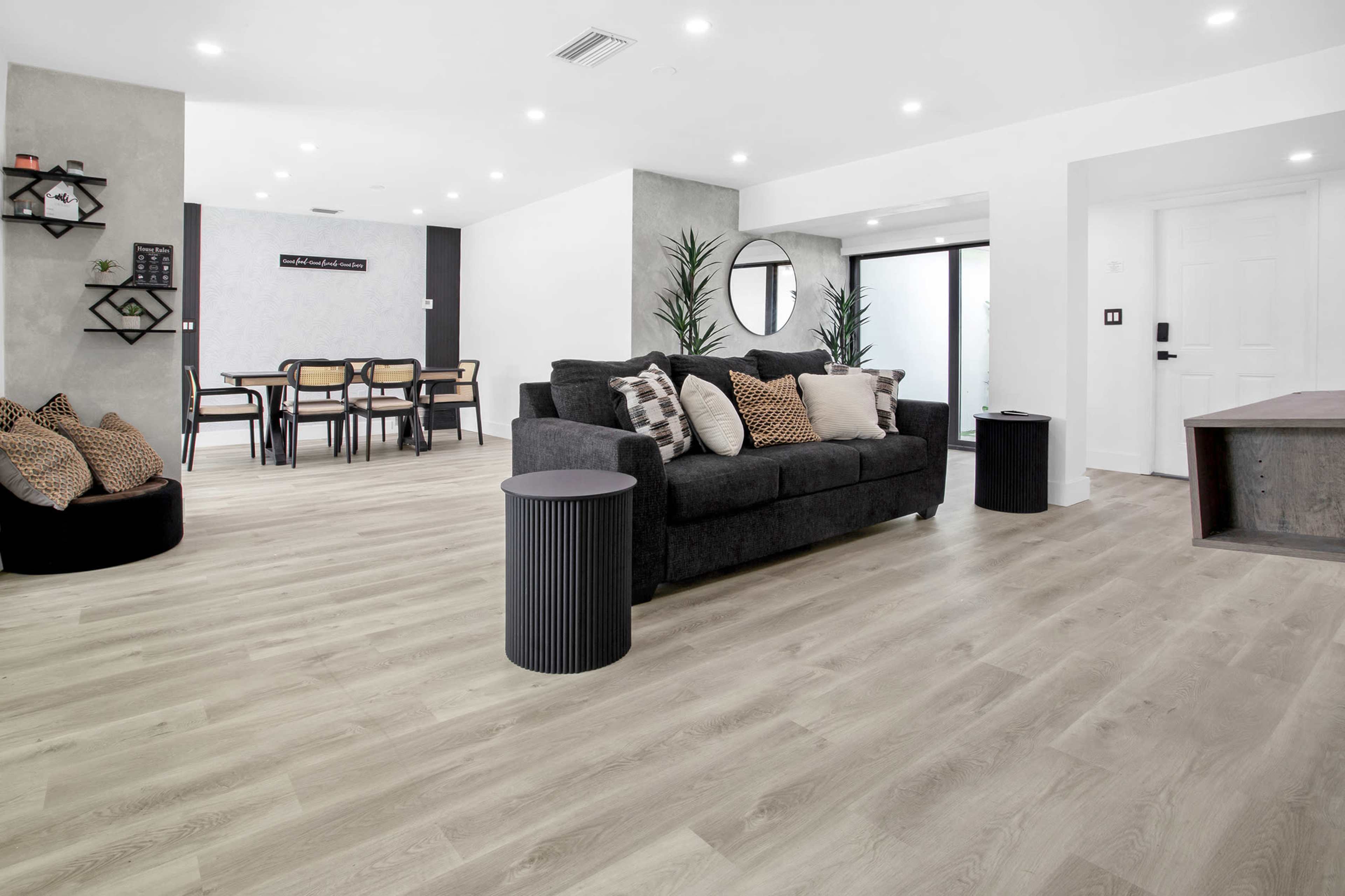 The image shows a modern interior living space featuring a dark sofa with cushions, a round side table, a dining area with chairs, and large windows providing natural light.