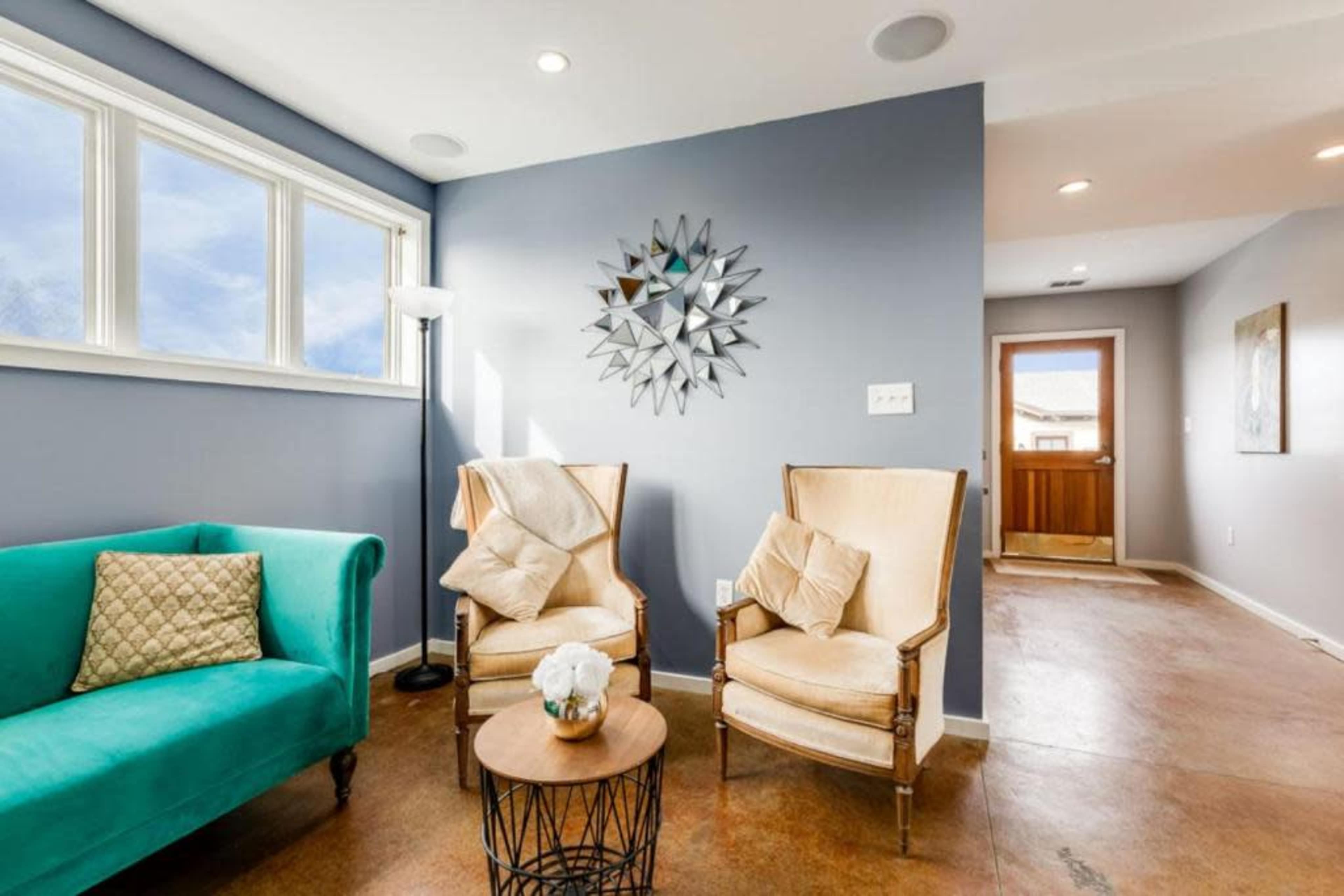 A cozy living area features a turquoise couch, two cream-colored armchairs, a circular wooden table, and a wall adorned with a decorative mirror.