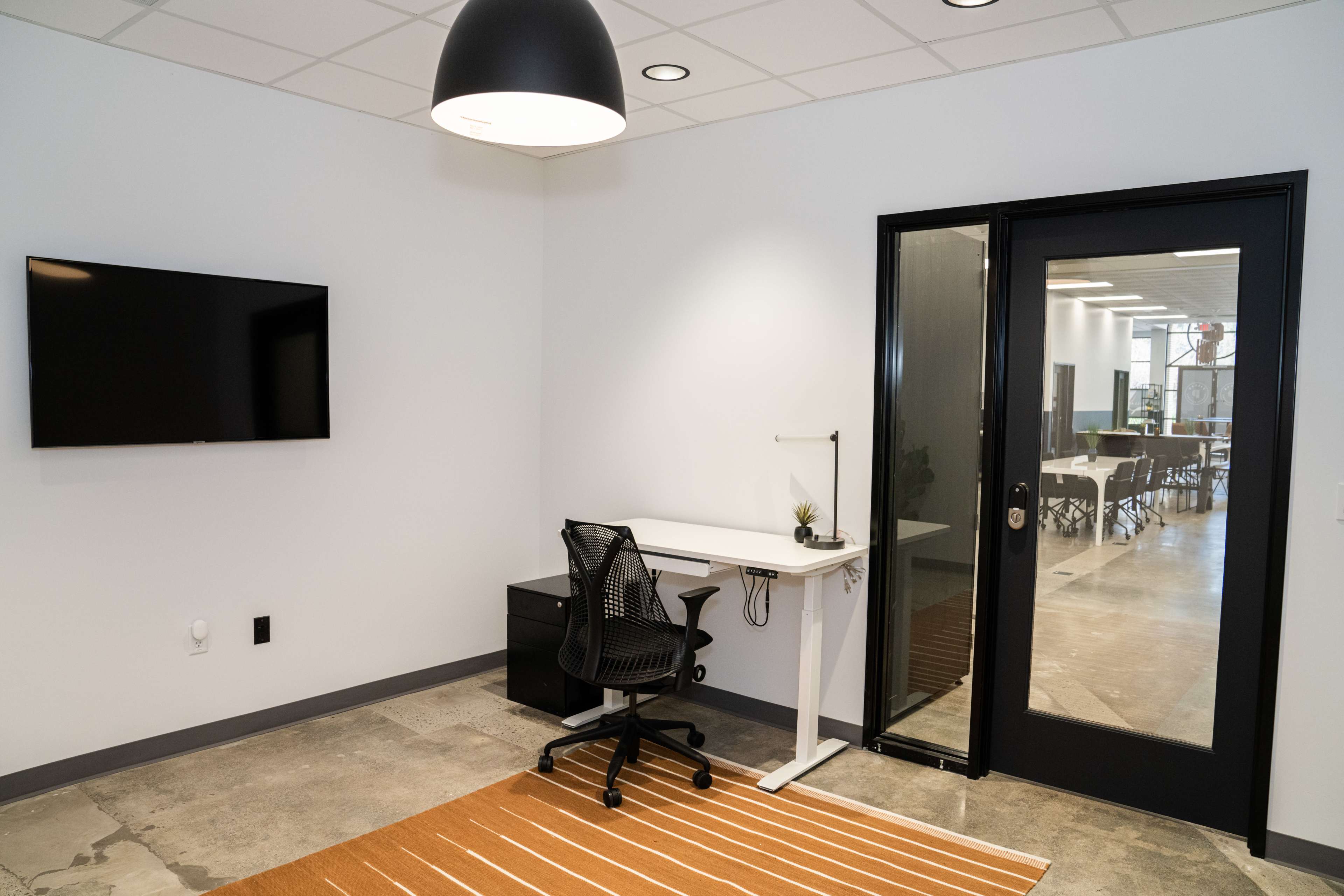 A modern office space featuring a desk with a chair, a wall-mounted television, and a glass door leading to an adjacent meeting area.