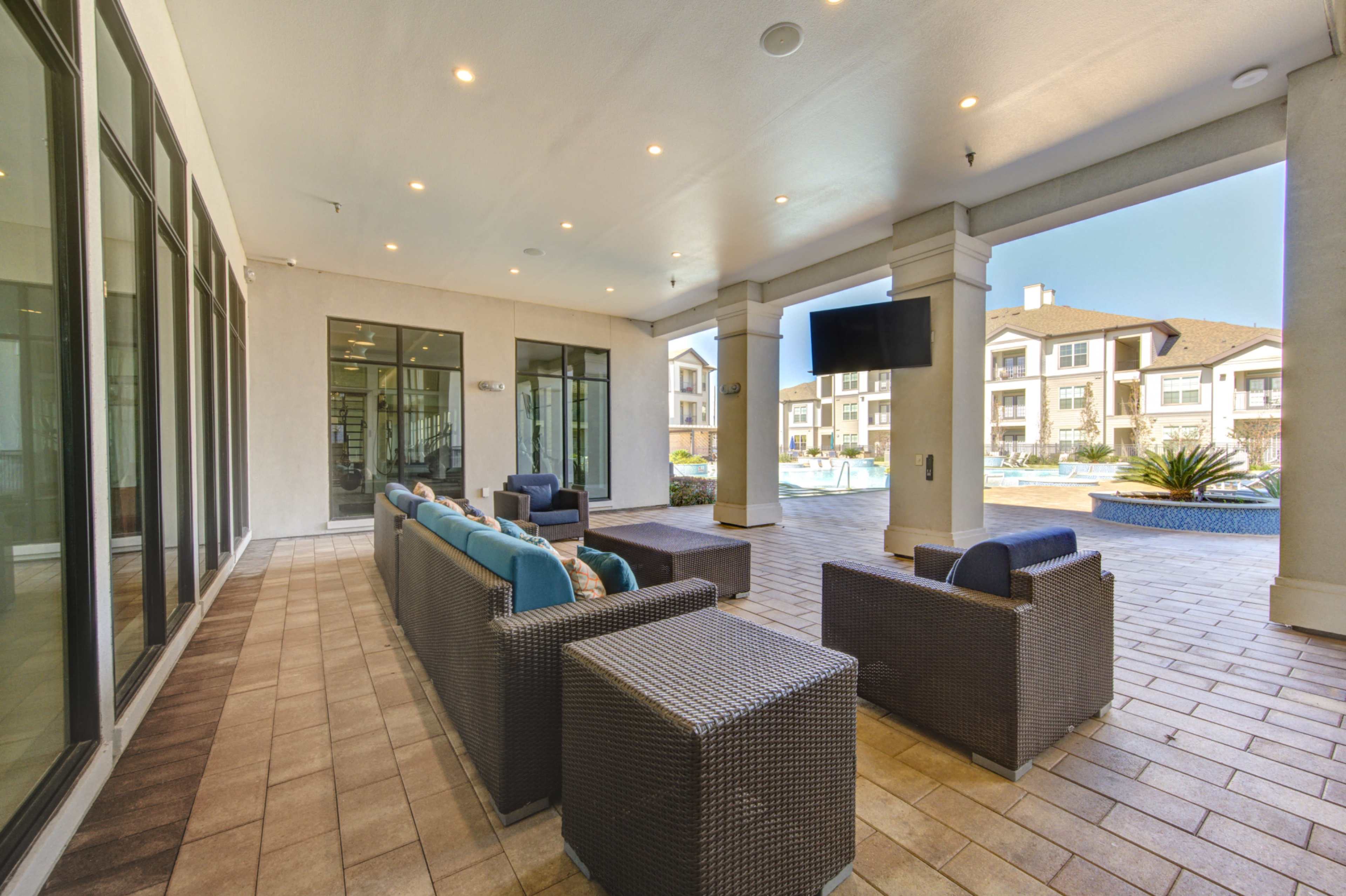 The image shows a covered outdoor lounge area with modern furniture, large windows, and a view of a pool and apartments beyond.
