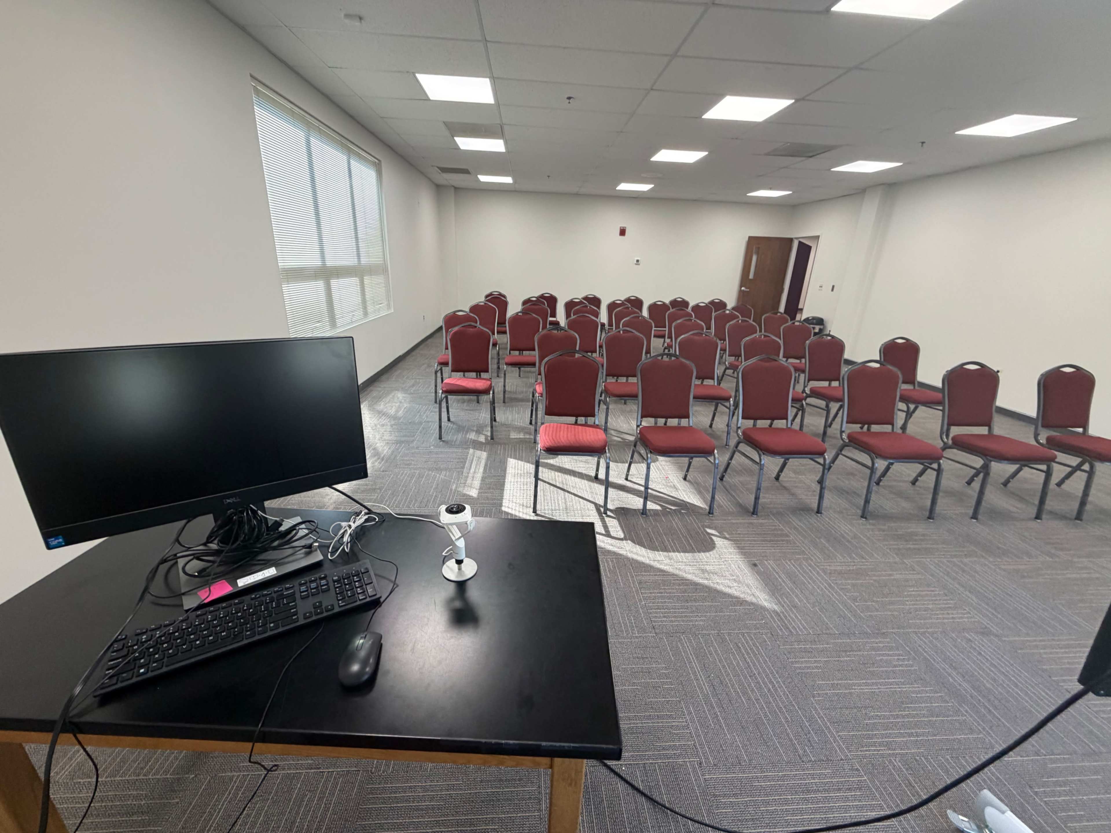 The image shows a room set up for a meeting or presentation, featuring a black table with a computer and webcam at the front, facing a row of empty red chairs arranged in neat rows.
