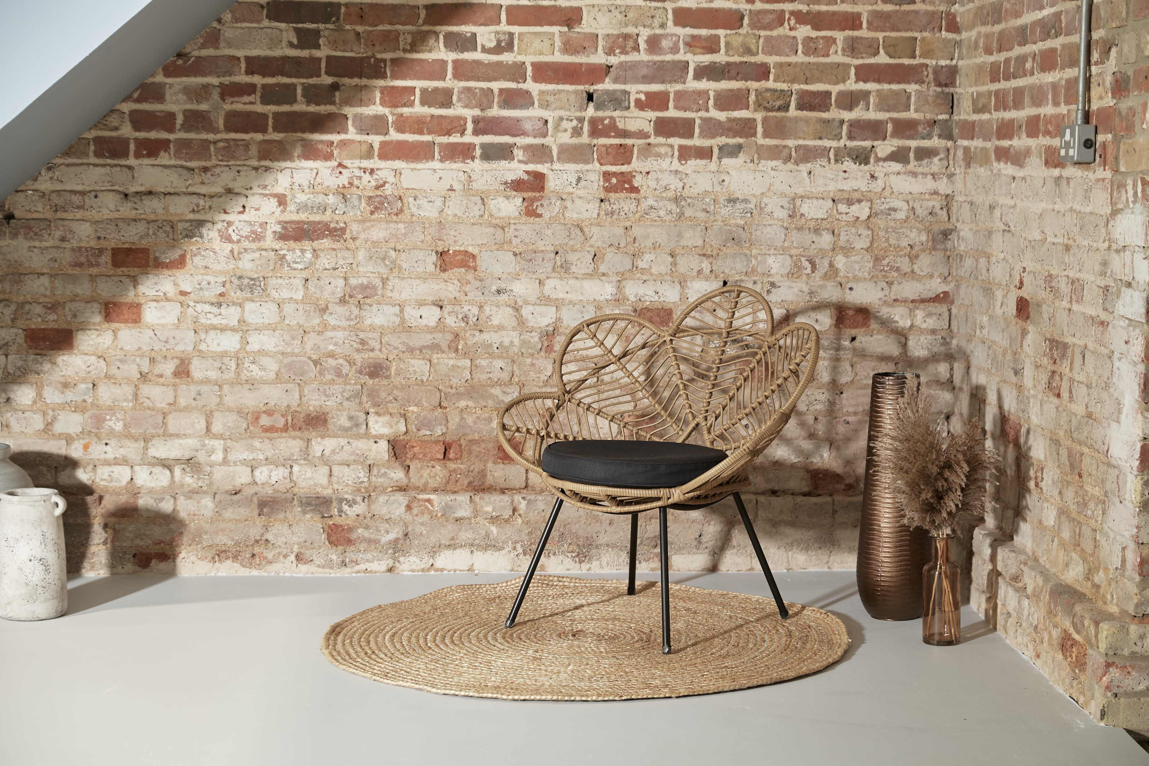 A woven rattan chair with a dark cushion sits on a round jute rug against a brick wall, accompanied by decorative vases and dried plants.