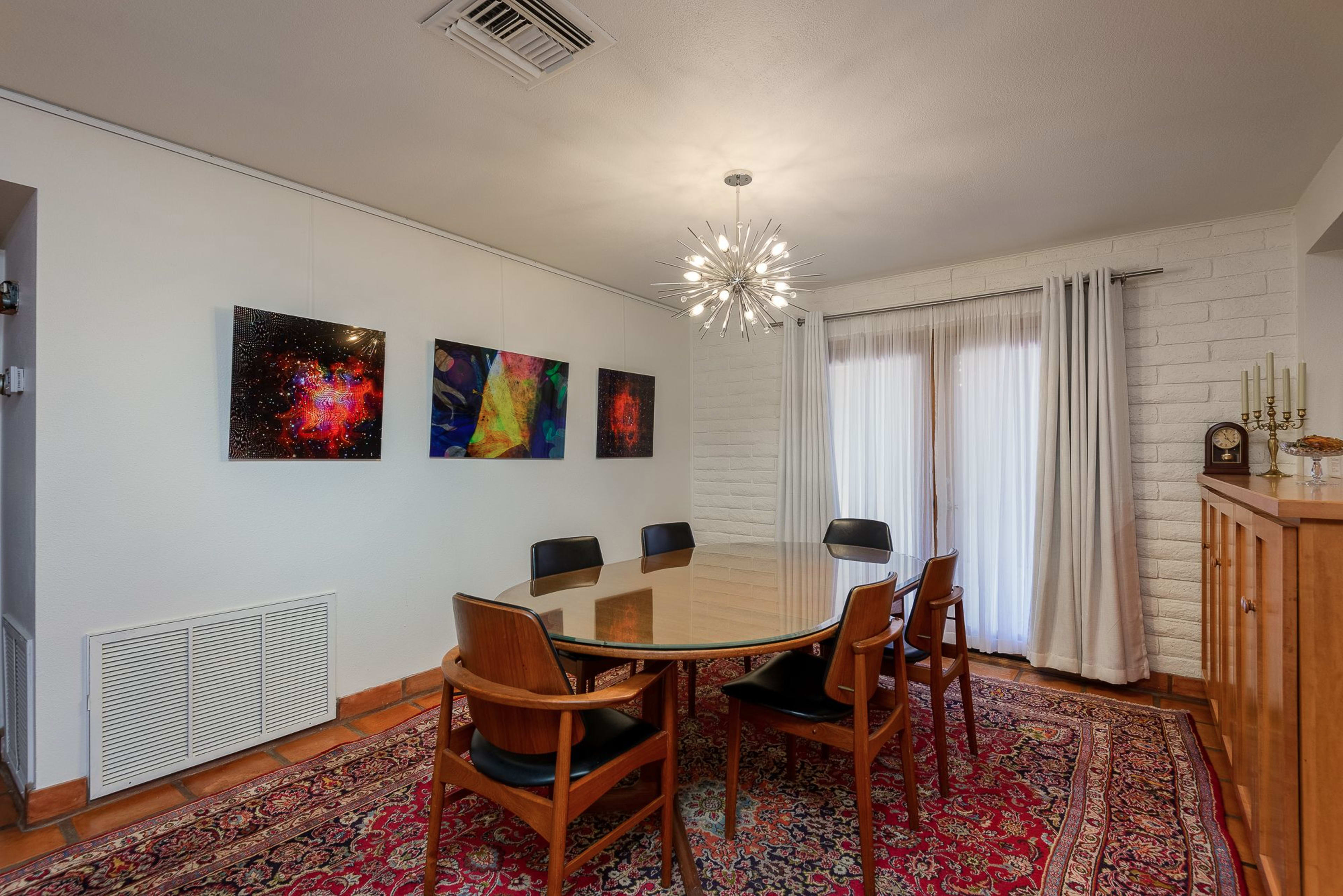 A modern dining room features a round glass table surrounded by wooden chairs, with colorful artwork on the walls and a light fixture overhead.
