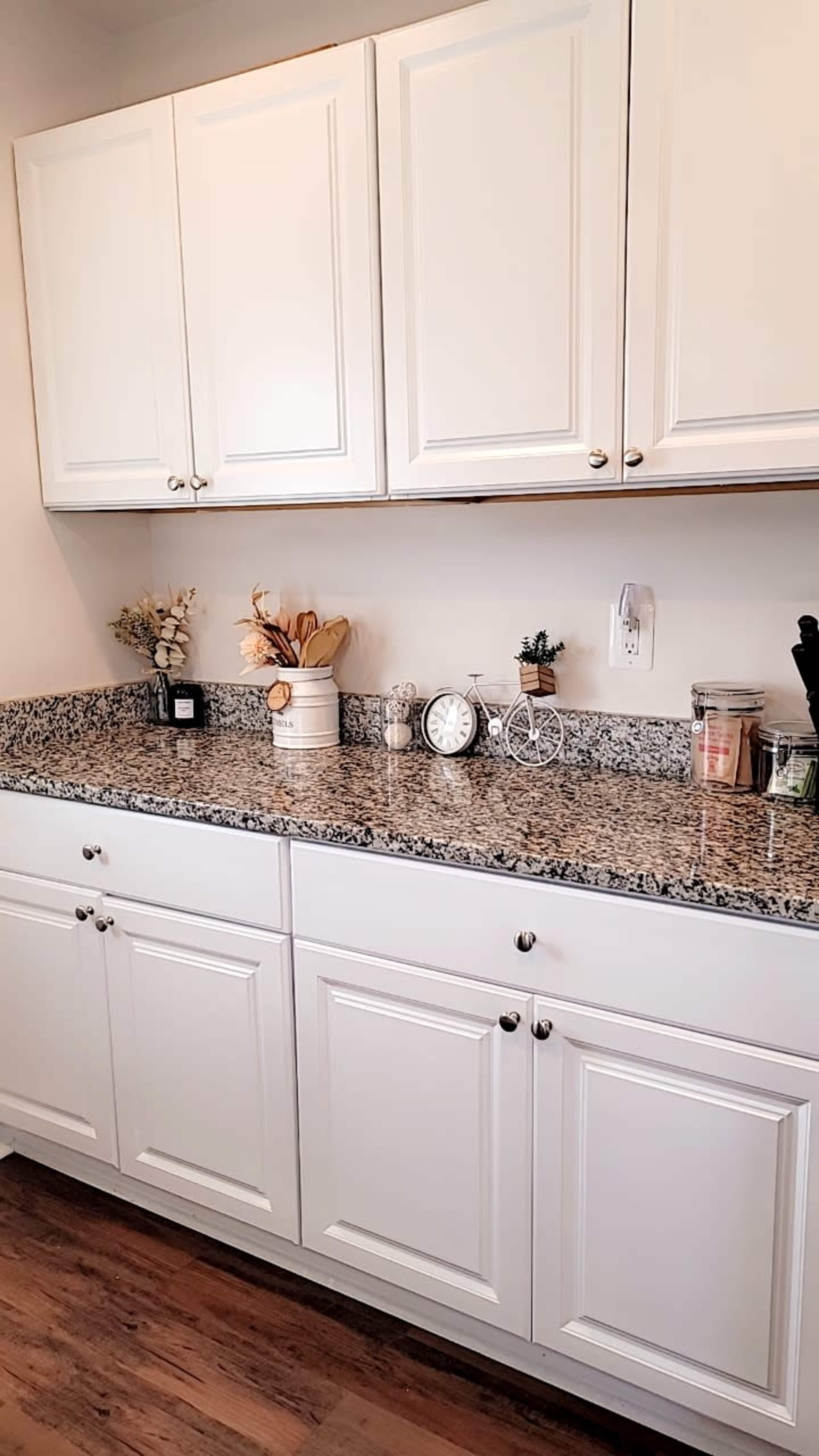 The image shows a kitchen countertop with granite surfaces, white cabinetry above, and various decorative items and kitchen tools arranged on the counter.