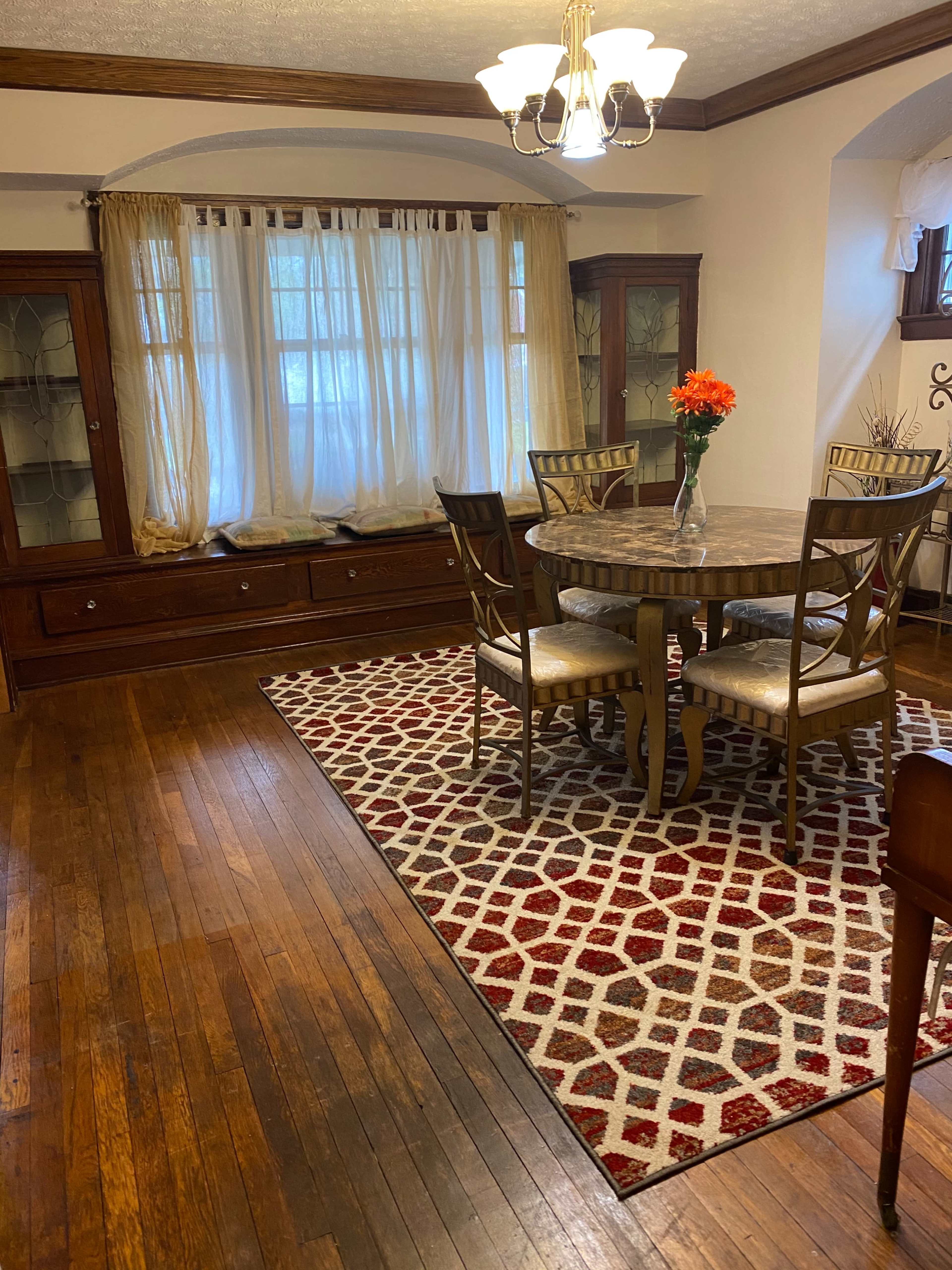 A dining area featuring a round table surrounded by four chairs, a floral centerpiece, a patterned rug, and large windows with light-colored drapes.