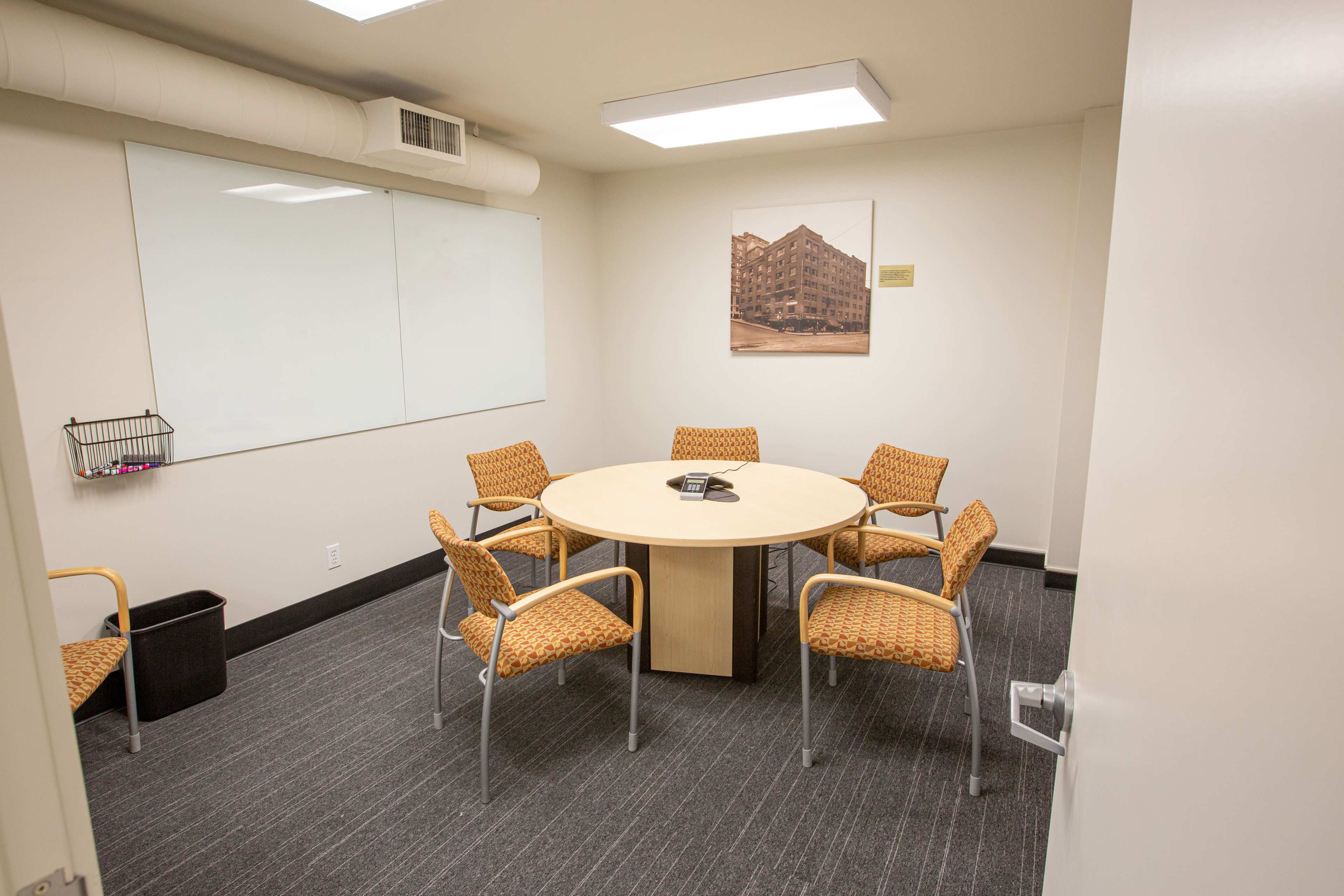 A small meeting room features a round table surrounded by four orange chairs, with a large whiteboard on one wall and a framed picture on another.