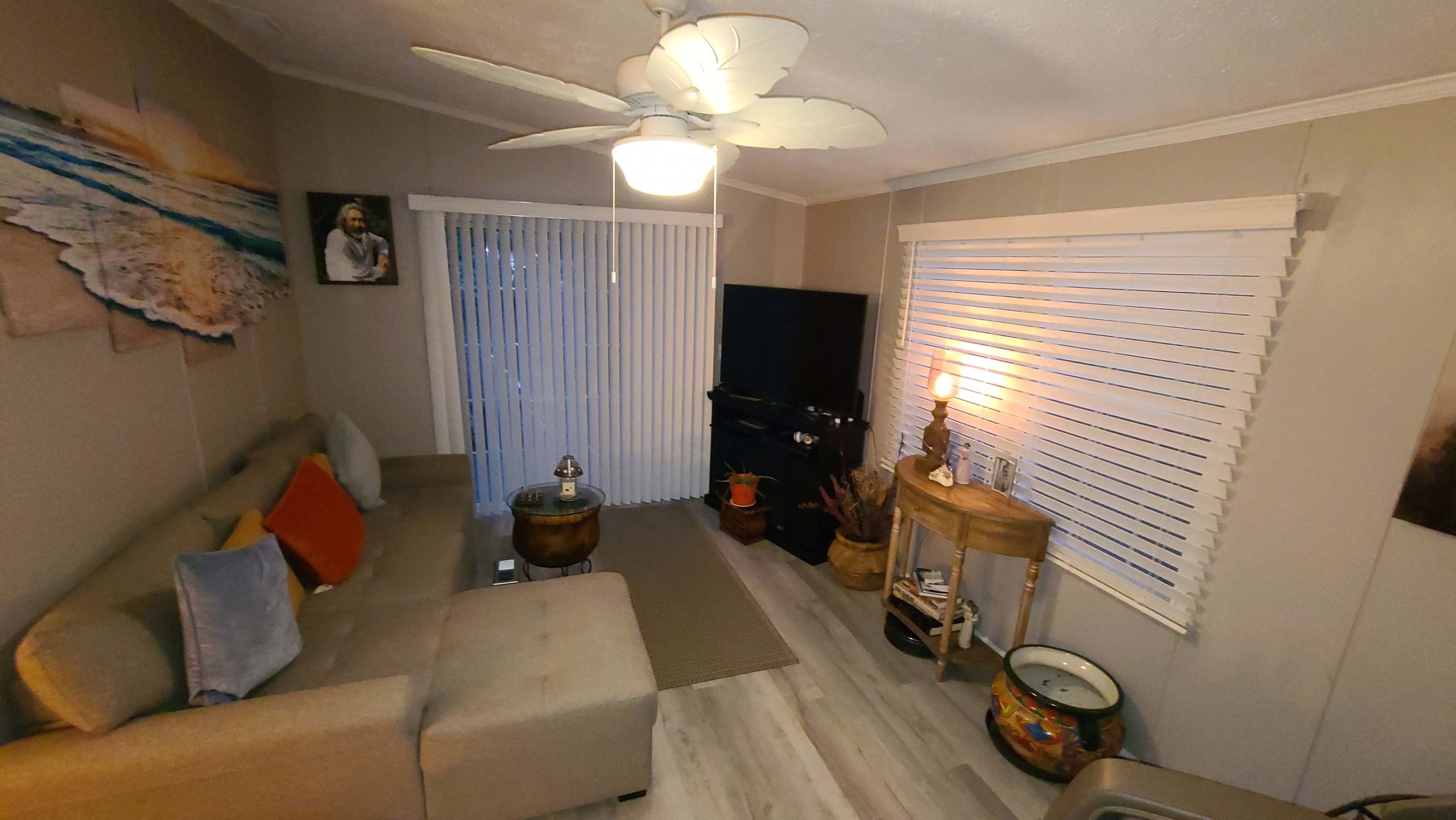 The living room features a sectional sofa with decorative pillows, a television on a black stand, and large windows covered with blinds, alongside a coffee table and a decorative bowl.