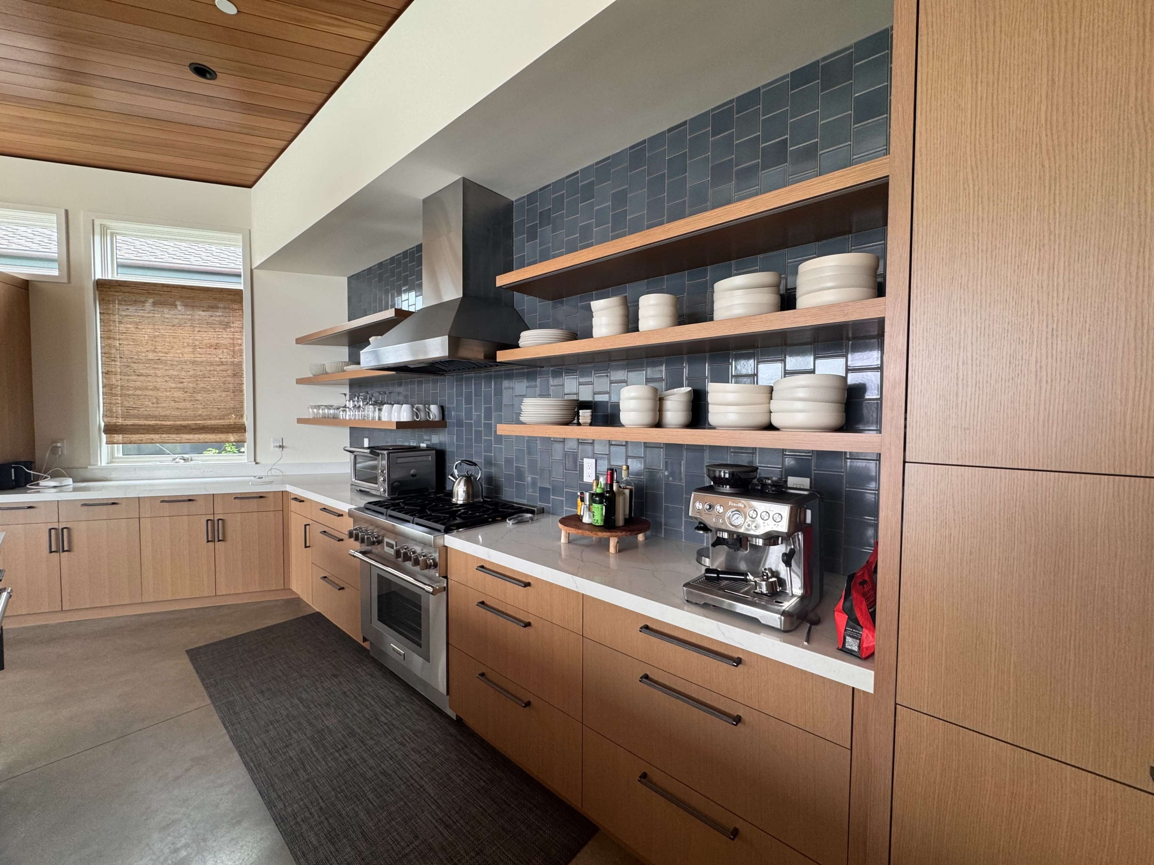 The image shows a modern kitchen with wooden cabinets, open shelves displaying ceramic dishes, and stainless steel appliances, including a gas range and a coffee machine.