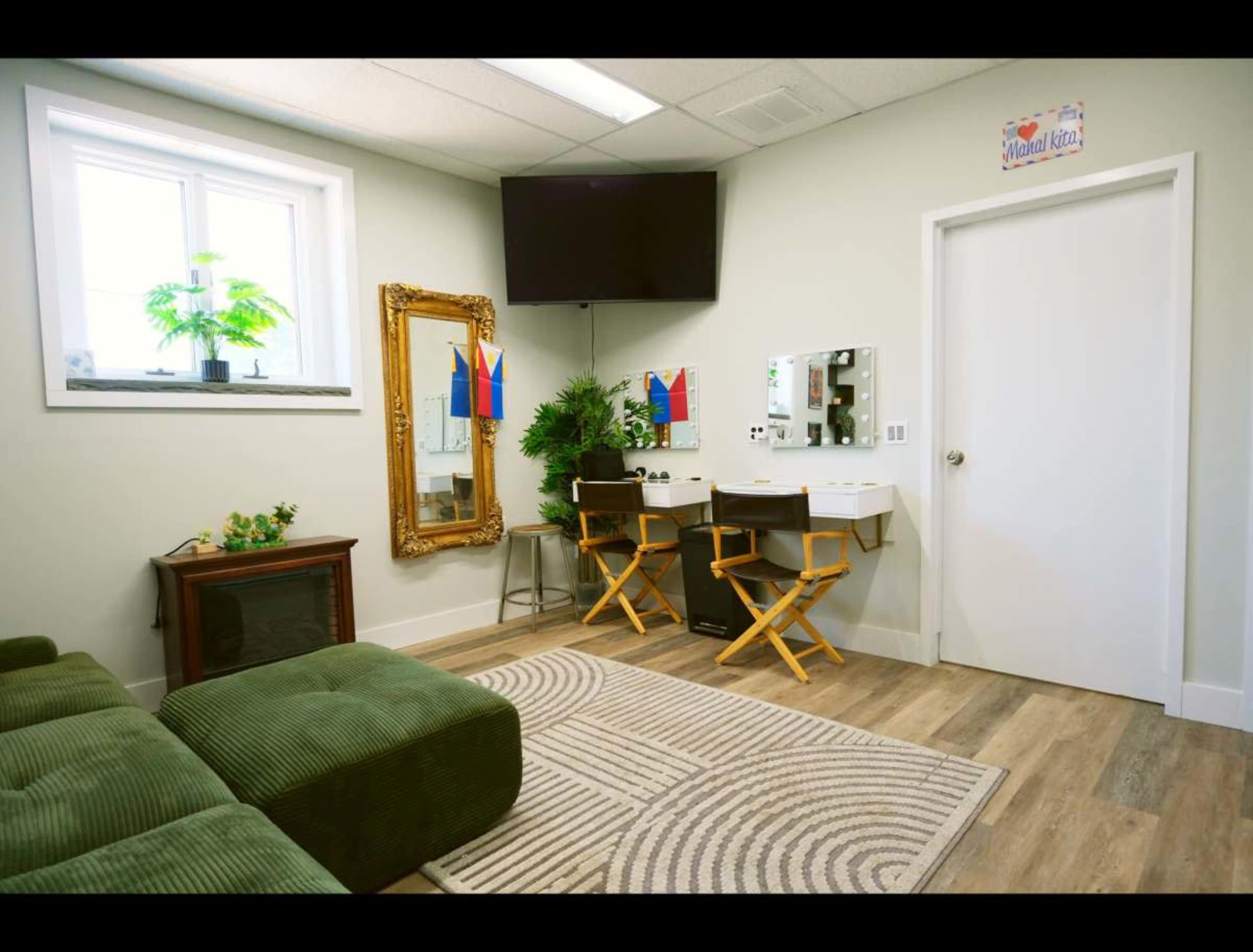 The image shows a cozy room with a green couch, a large wall mirror with lights, two director's chairs, a television, and plants near a window.