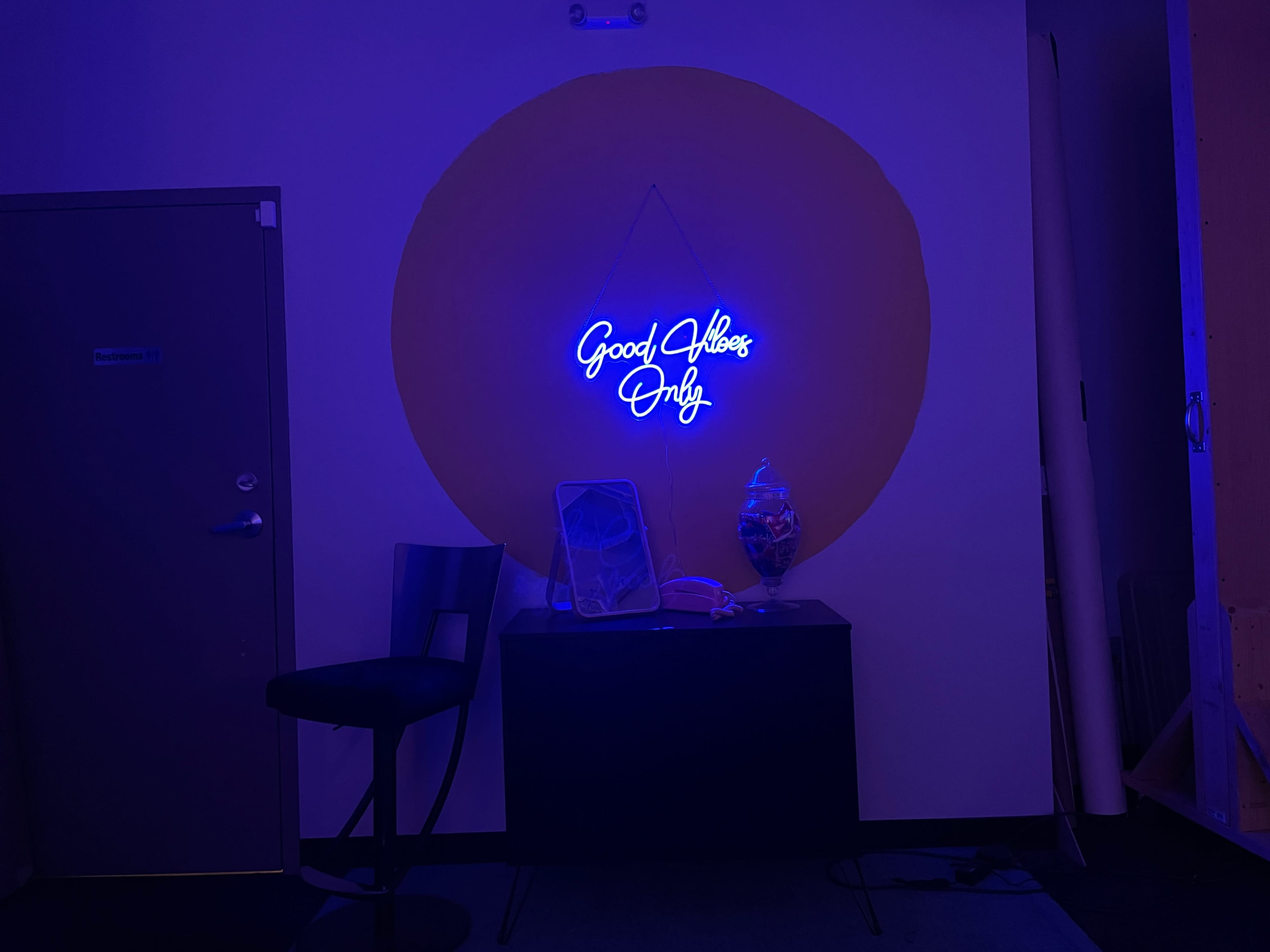 A blue neon sign reading "Good Vibes Only" is illuminated on a wall above a dark cabinet, accompanied by a chair and a decorative glass object.