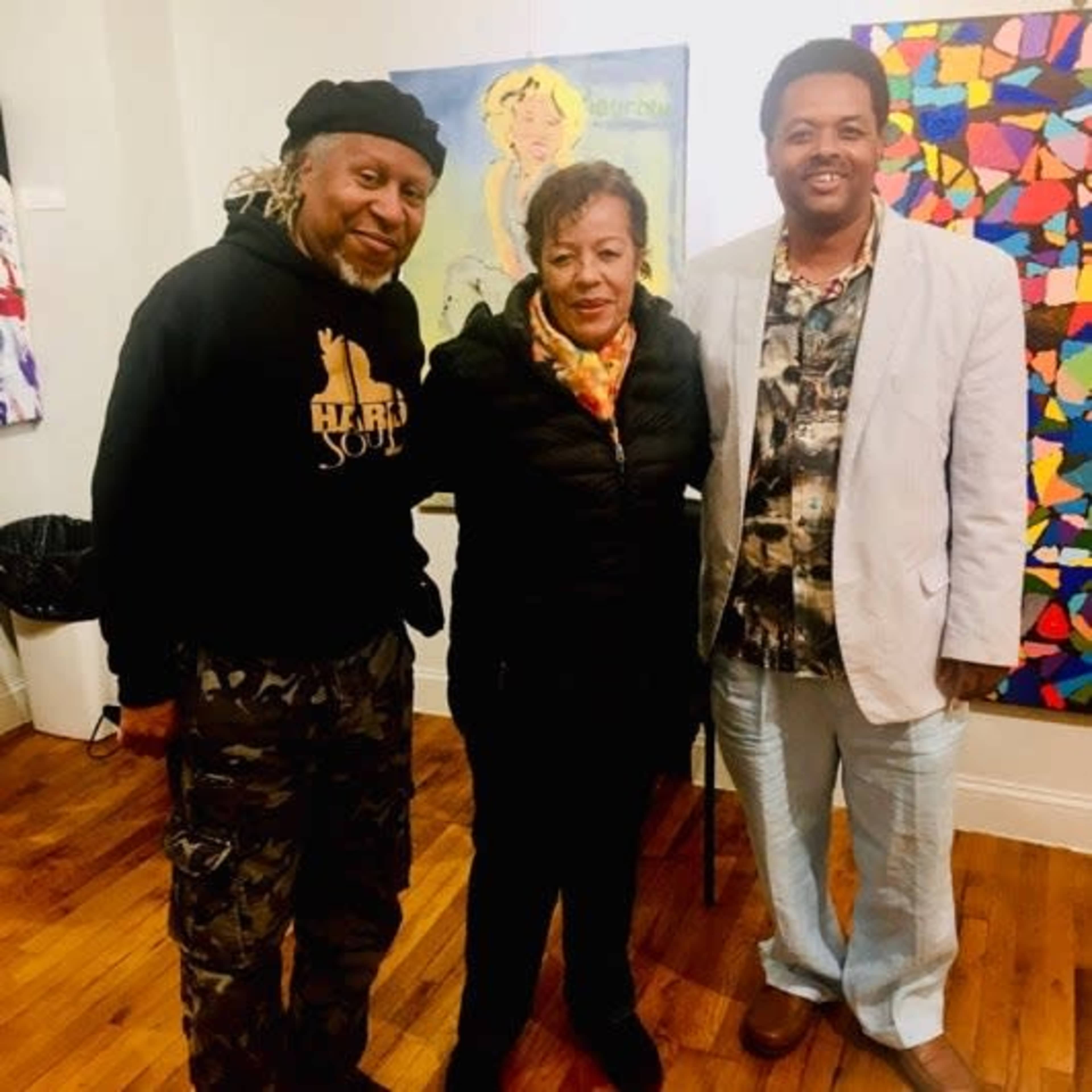 Three individuals stand together in an art gallery, with colorful artworks visible in the background.