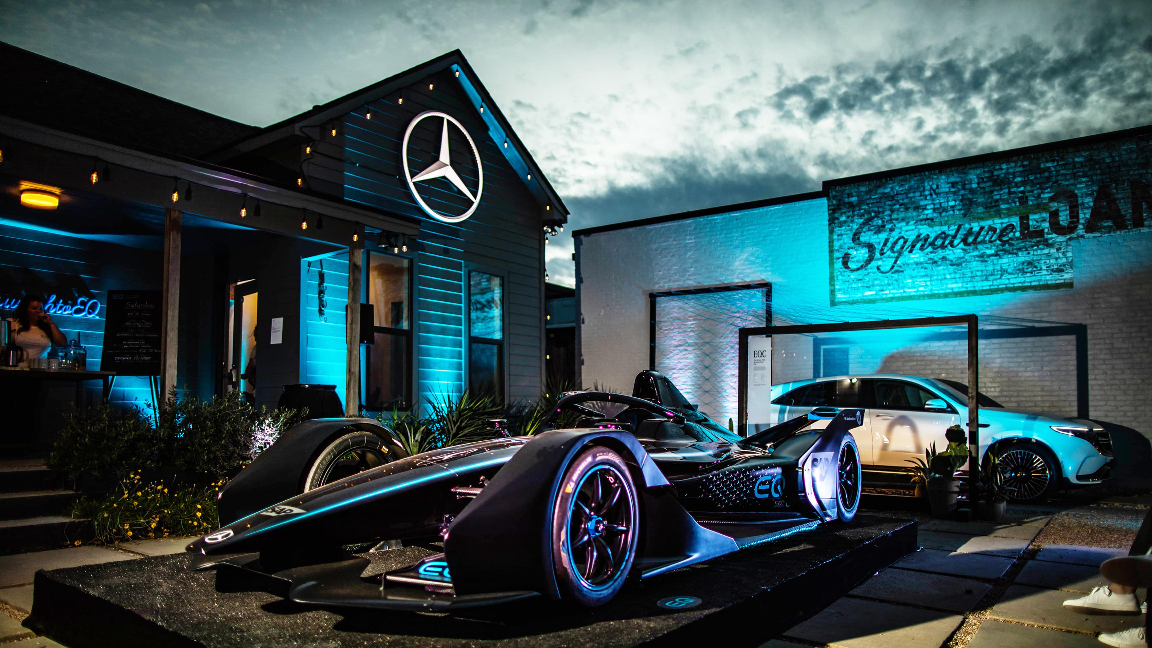 A Mercedes-Benz racing car is prominently displayed outside a building adorned with the Mercedes logo and blue lighting.