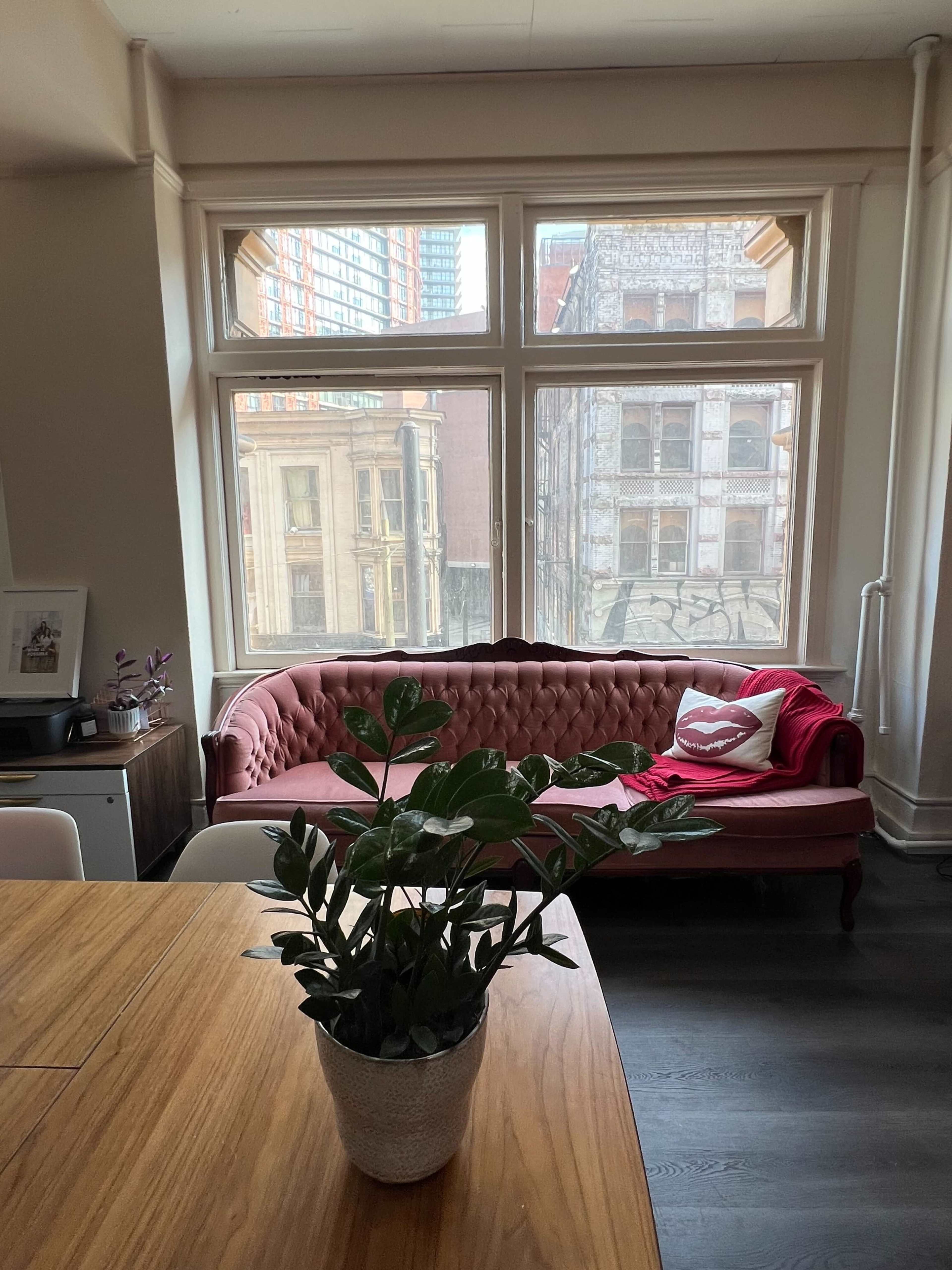 A cozy living room features a pink tufted sofa, a wooden dining table, and a potted plant, with large windows showing a city view.