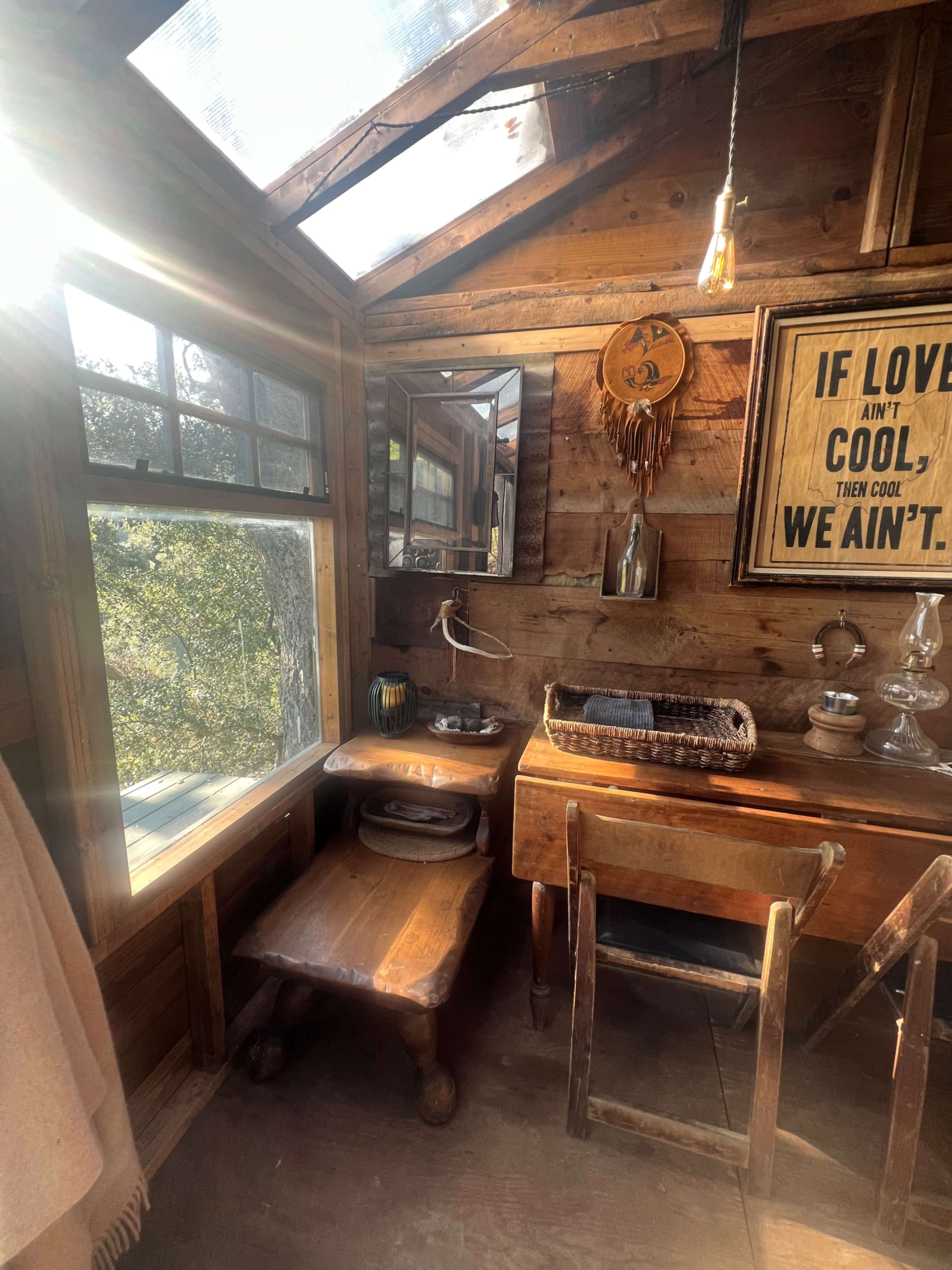 The image shows a rustic wooden interior with a small desk, a chair, a mirror, and a framed sign on the wall.