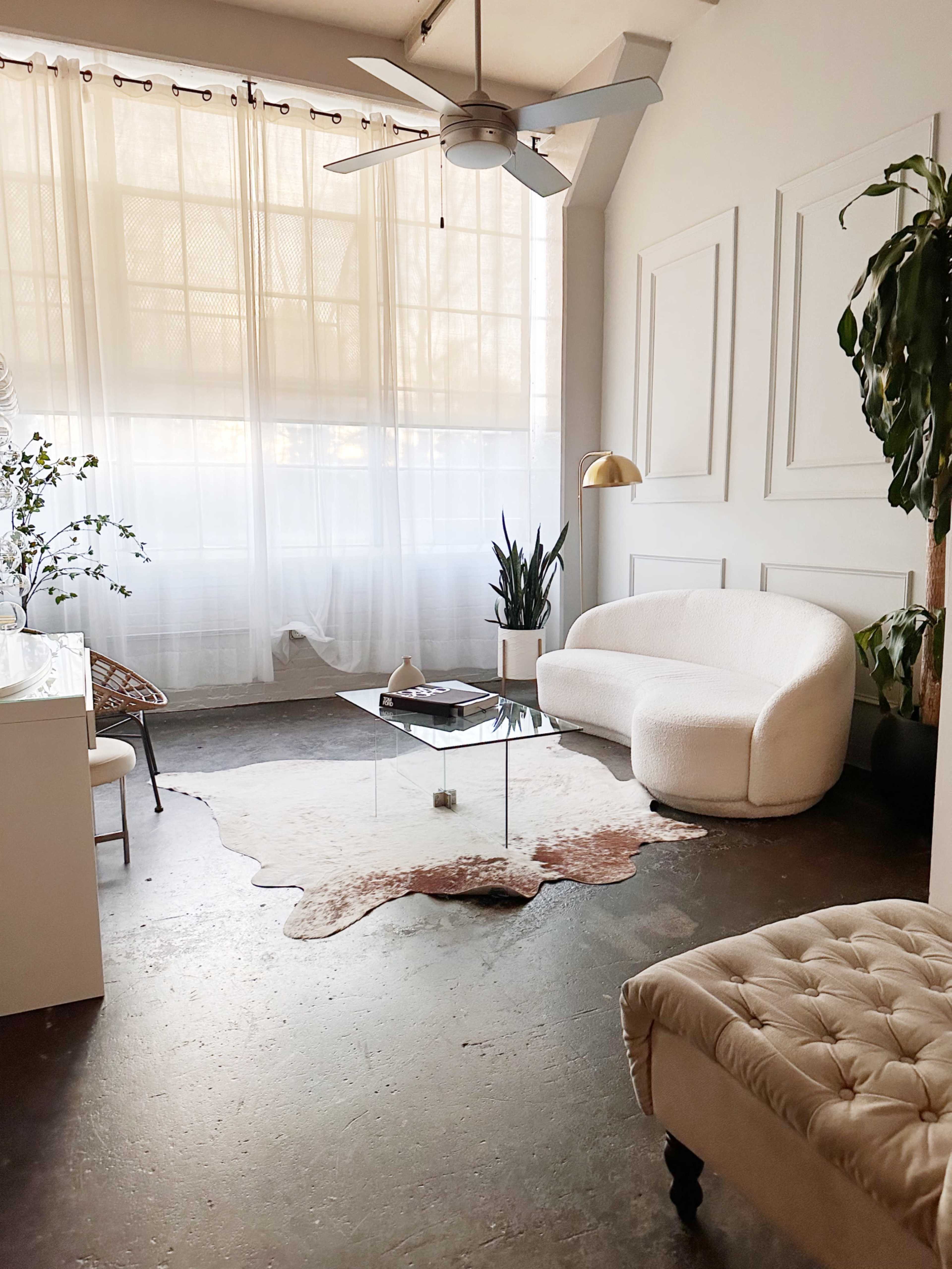 A modern living room with a curved white sofa, a glass coffee table on a patterned rug, and large windows draped with sheer curtains.