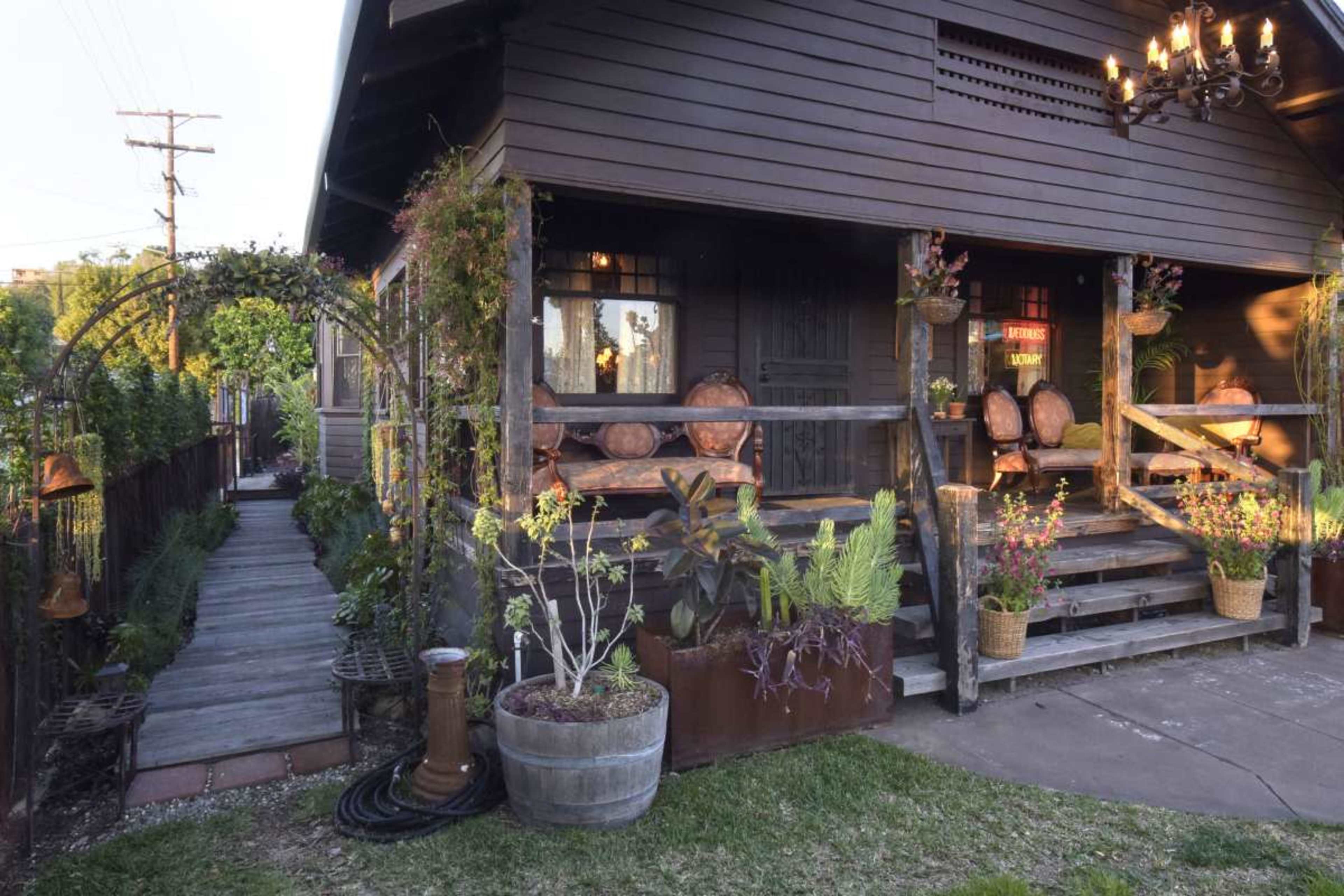 The image shows a wooden house with a porch adorned with potted plants and decorative elements, along with a winding pathway leading to the entrance.