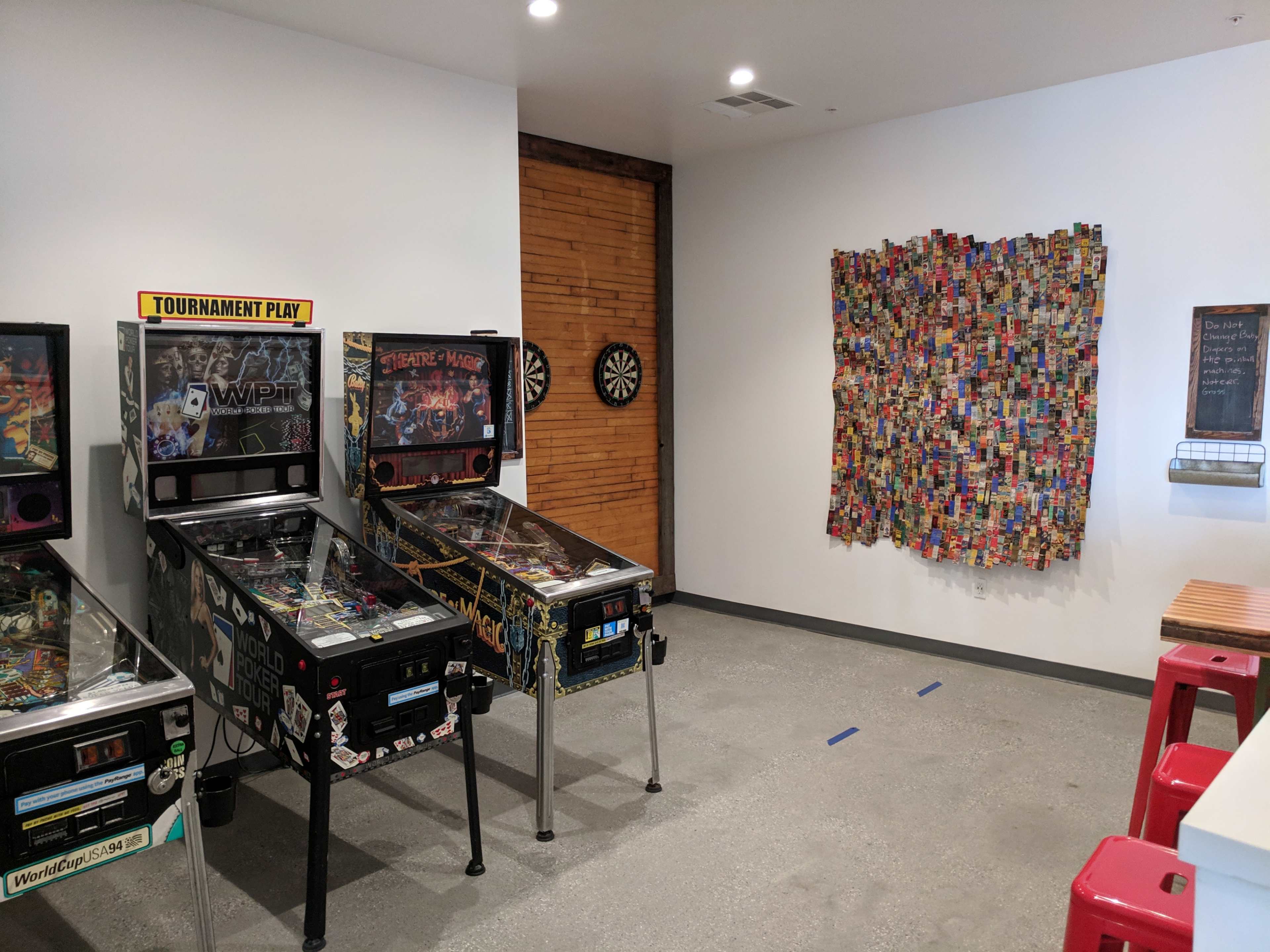The image shows a game room with three pinball machines on one side and a colorful piece of wall art made from various items on the opposite wall.
