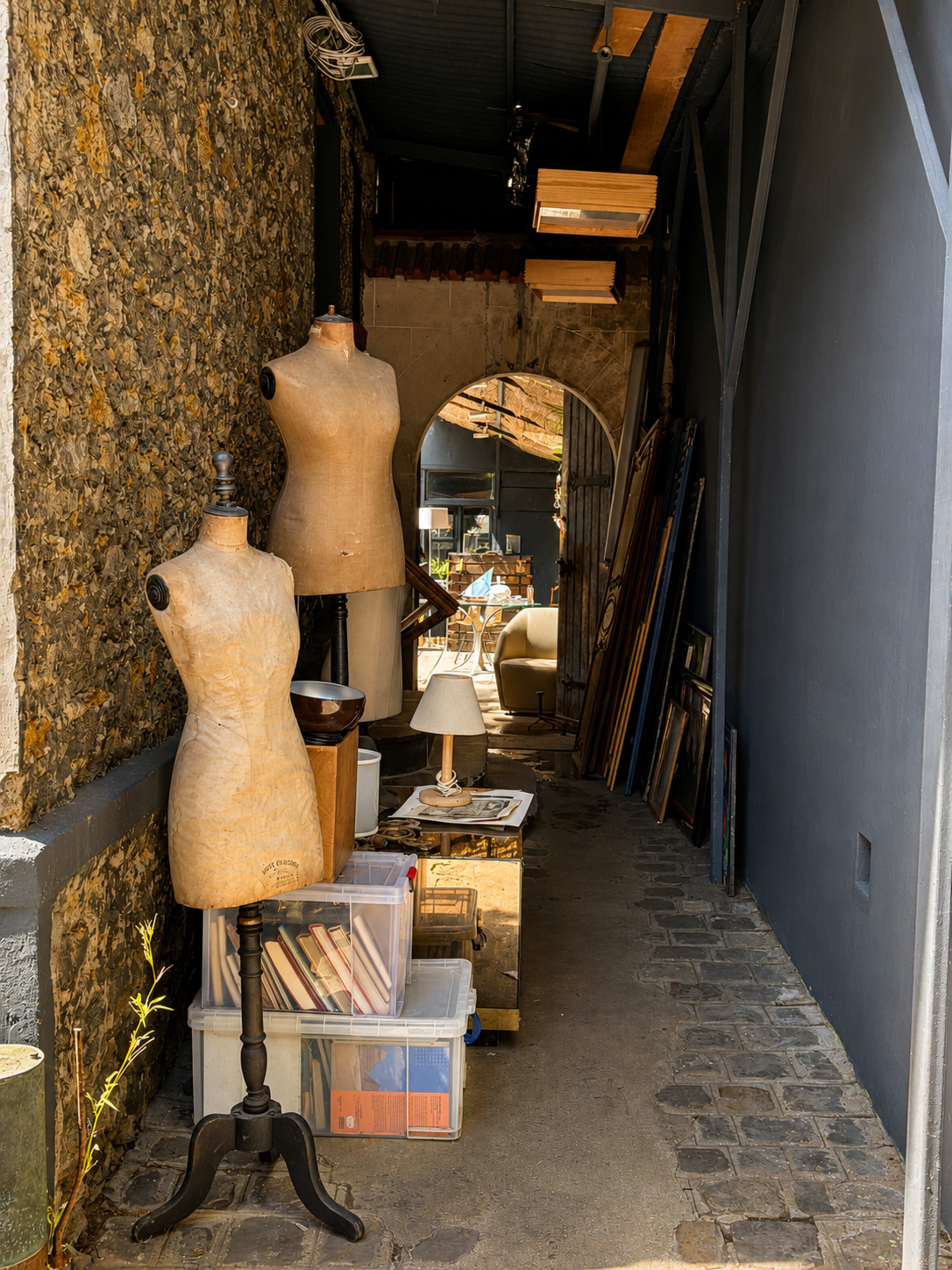The image shows a narrow alleyway with two dressmaker's mannequins on display alongside various boxes and art items.