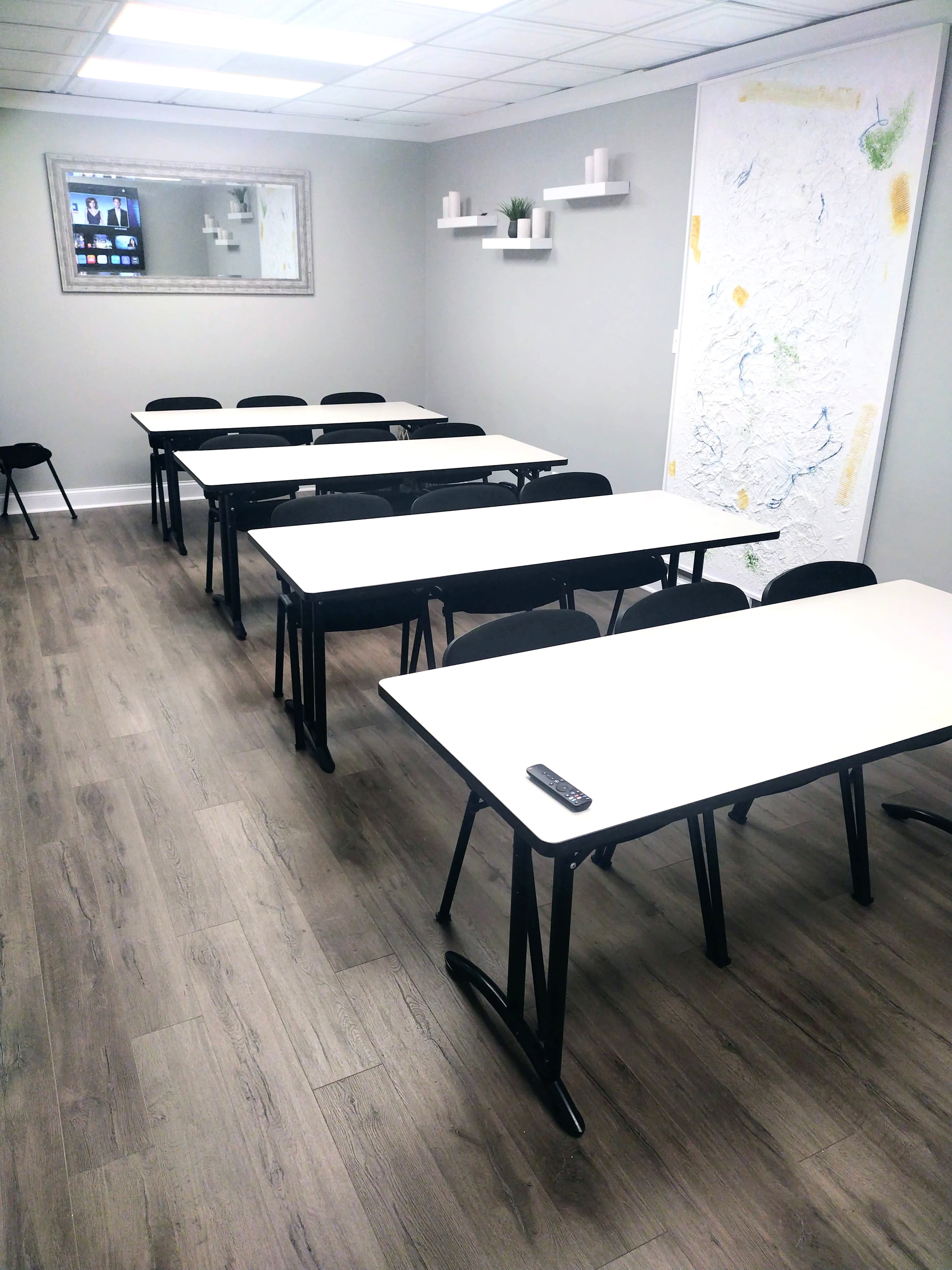 A classroom with several black chairs and white tables arranged in rows along a wooden floor, featuring a wall with a mirror and a decorative map.