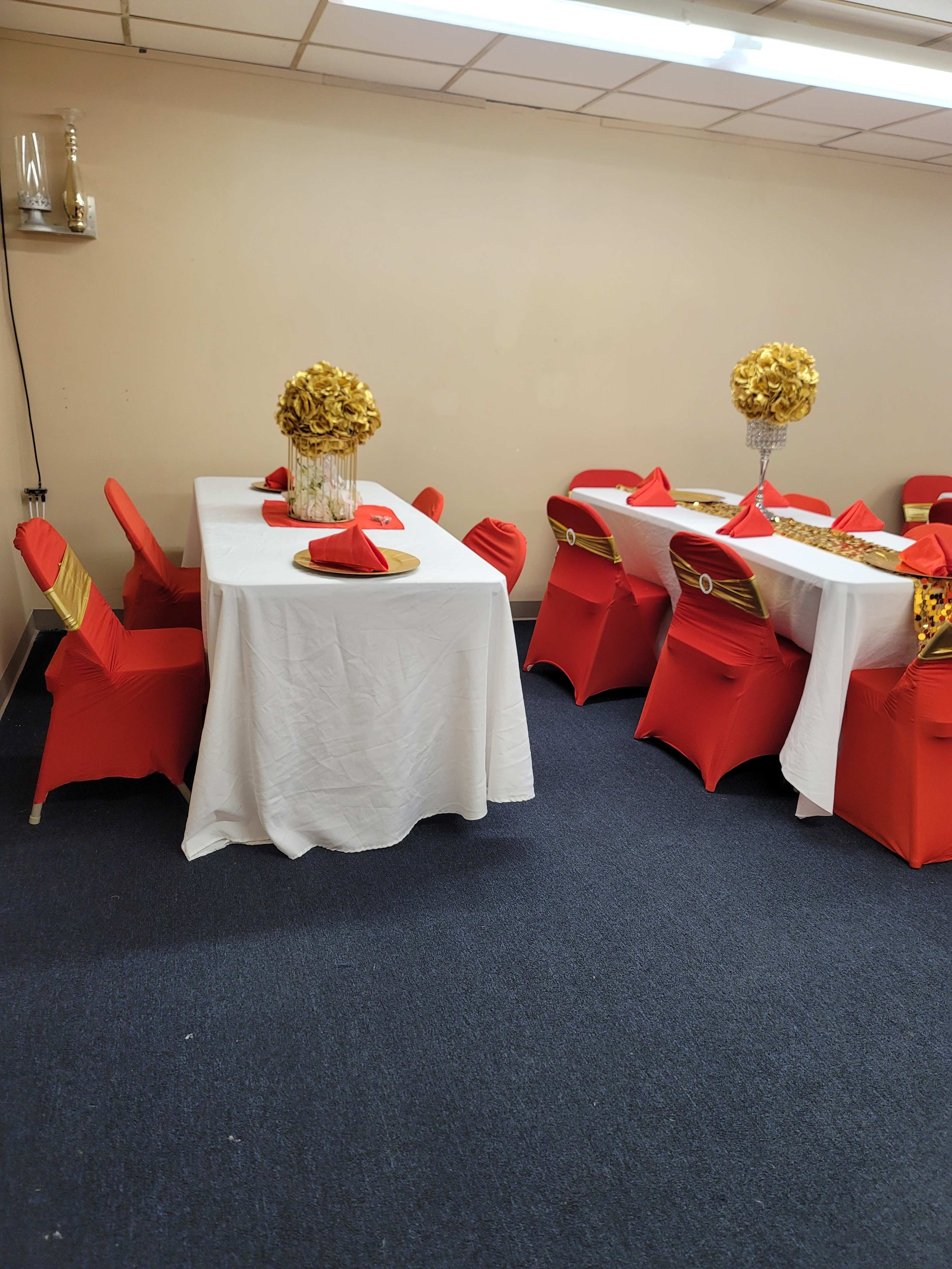 A dining area features two tables covered in white tablecloths with red chairs adorned with gold sashes and decorative centerpieces.