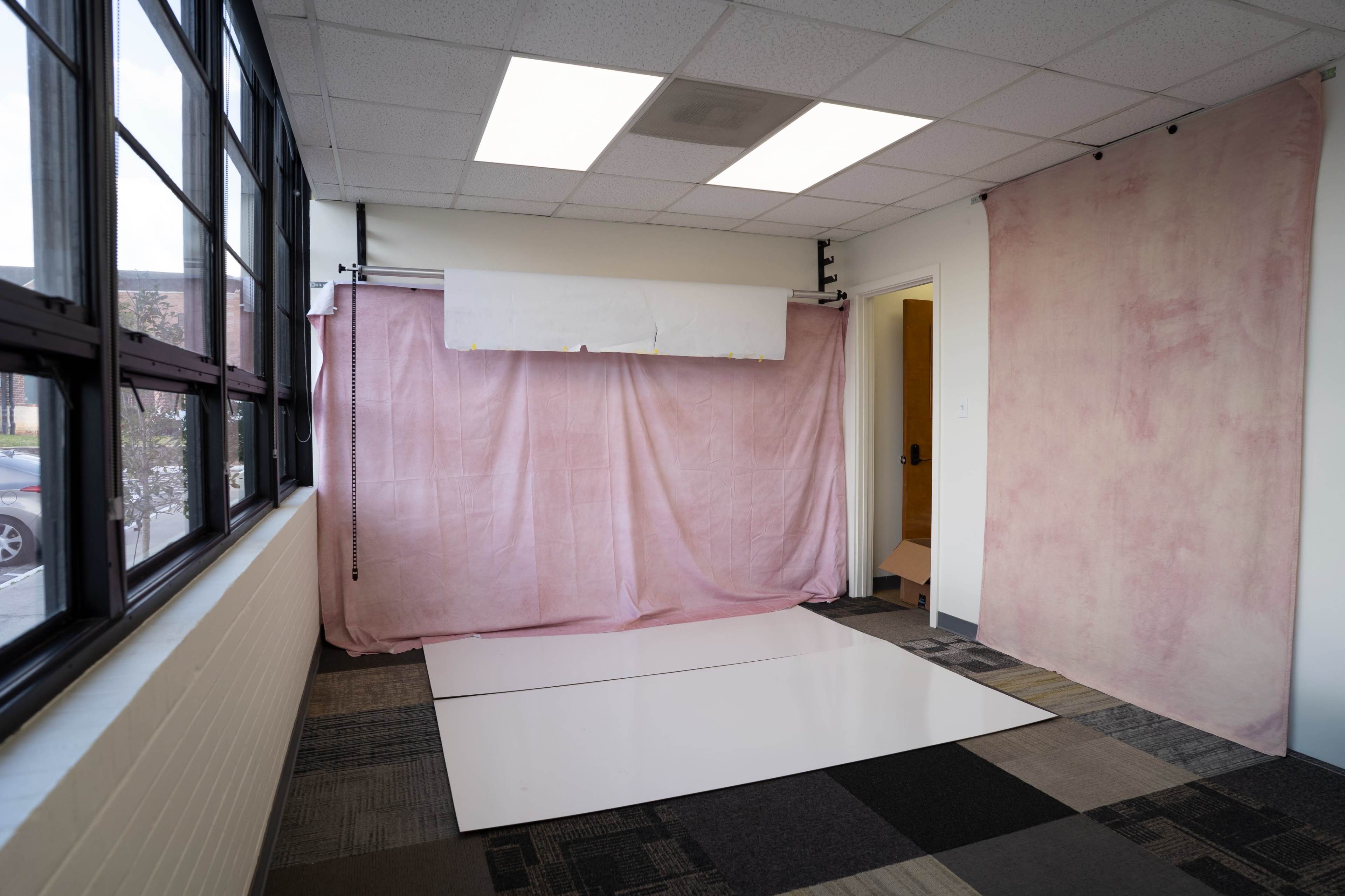 The image shows a photography studio with a pink backdrop, white floor, and large windows letting in natural light.