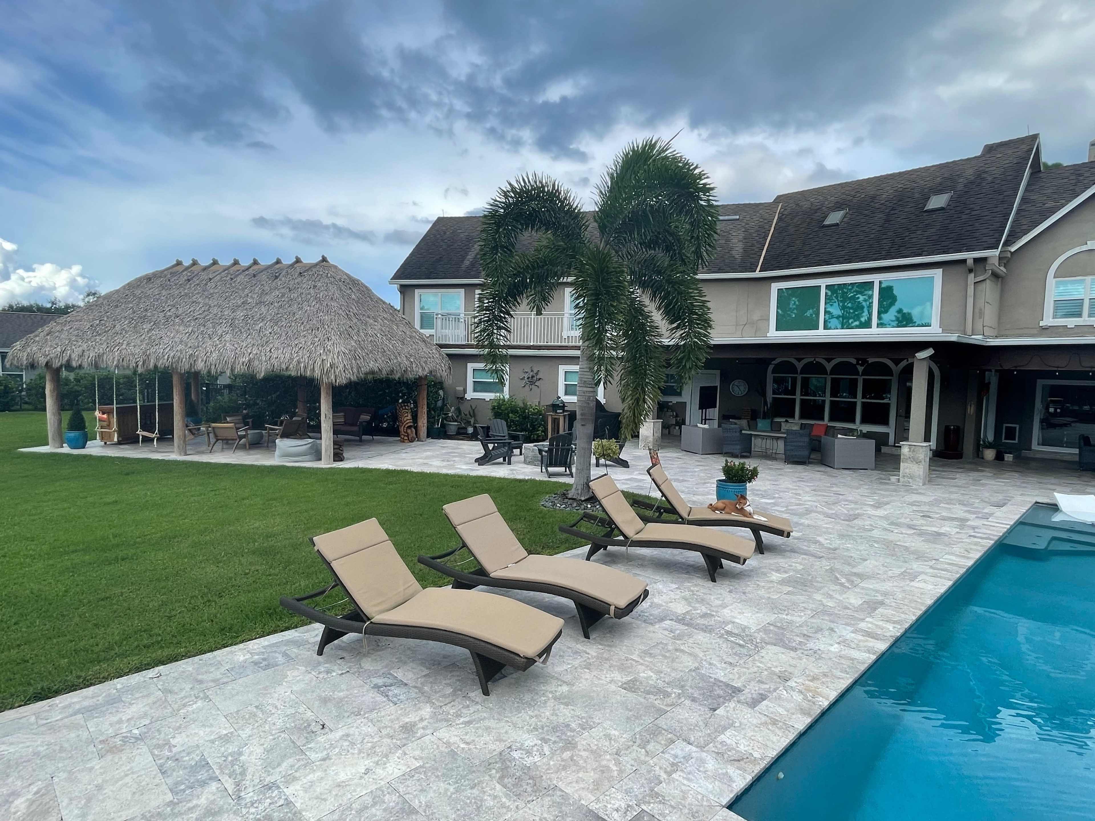 A backyard features a swimming pool, loungers, and a thatched gazebo surrounded by a well-maintained lawn and a spacious house.